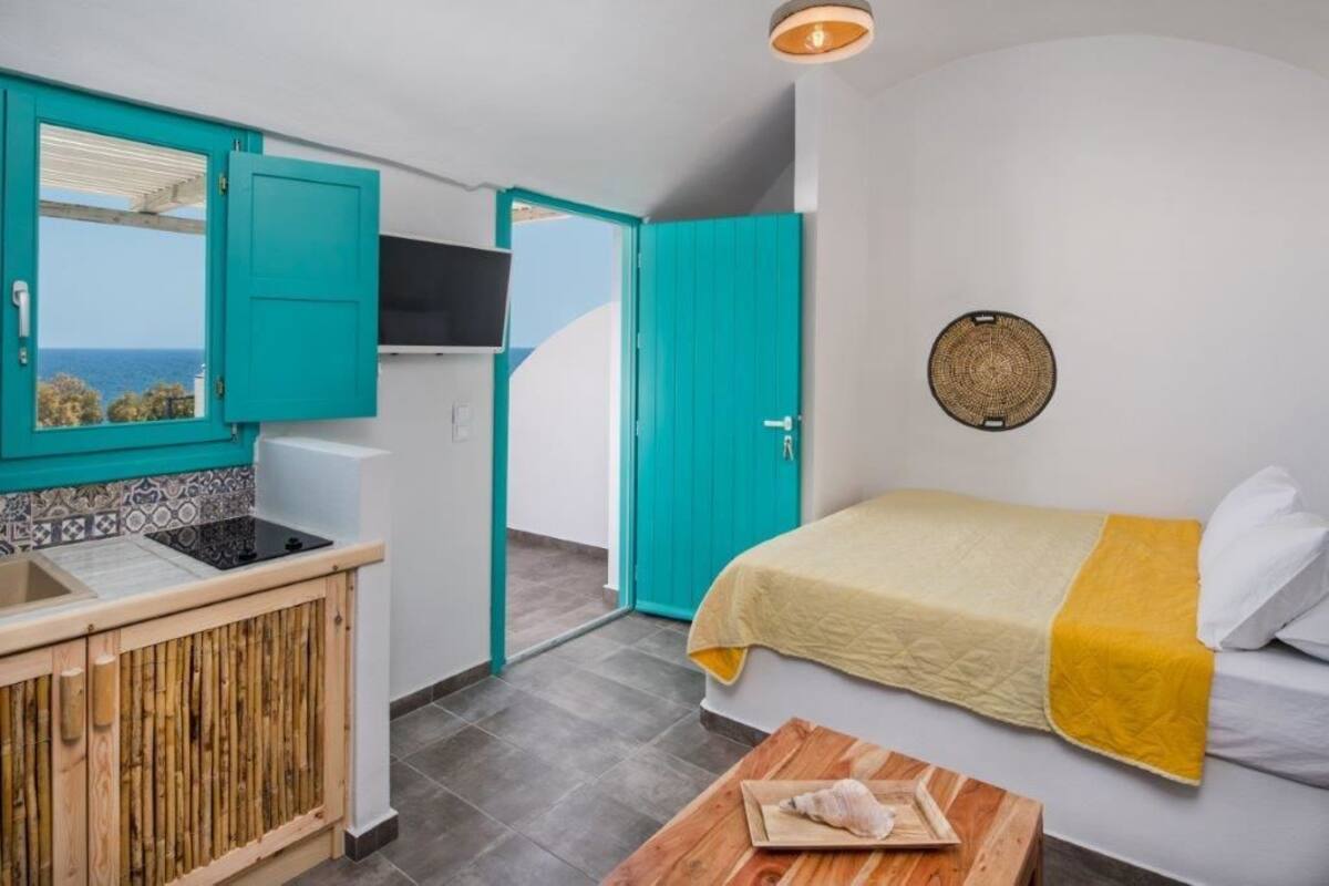 A compact room with a kitchenette, a bed with a yellow blanket, and a view of the sea through an open door.