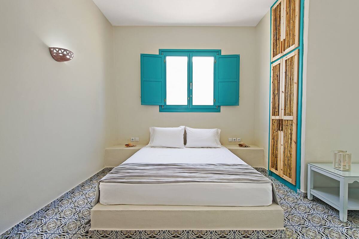 A bedroom with a bed, two nightstands, a wall-mounted light, and a window with blue shutters.