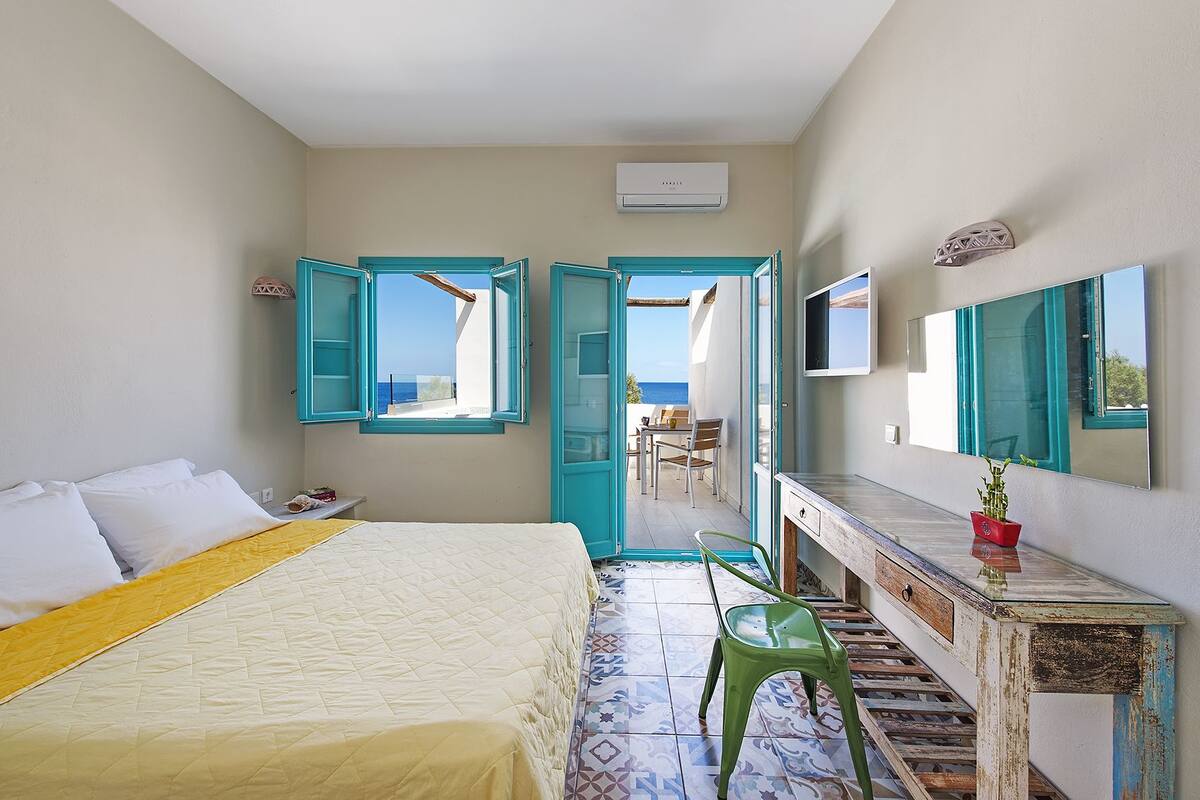 A bedroom with a bed, a desk, a chair, and a view of the sea through open doors.