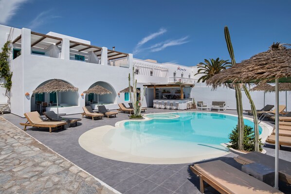 A pool area with lounge chairs, a bar, and a building with white walls and arches. - Nissia Beach Apartments and Suites (Santorini)