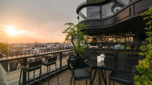 Un bar situé sur le toit d’un immeuble, offrant une vue sur la ville, avec une rambarde en verre incurvée, un espace bar où sont exposées les bouteilles et des sièges en plein air.