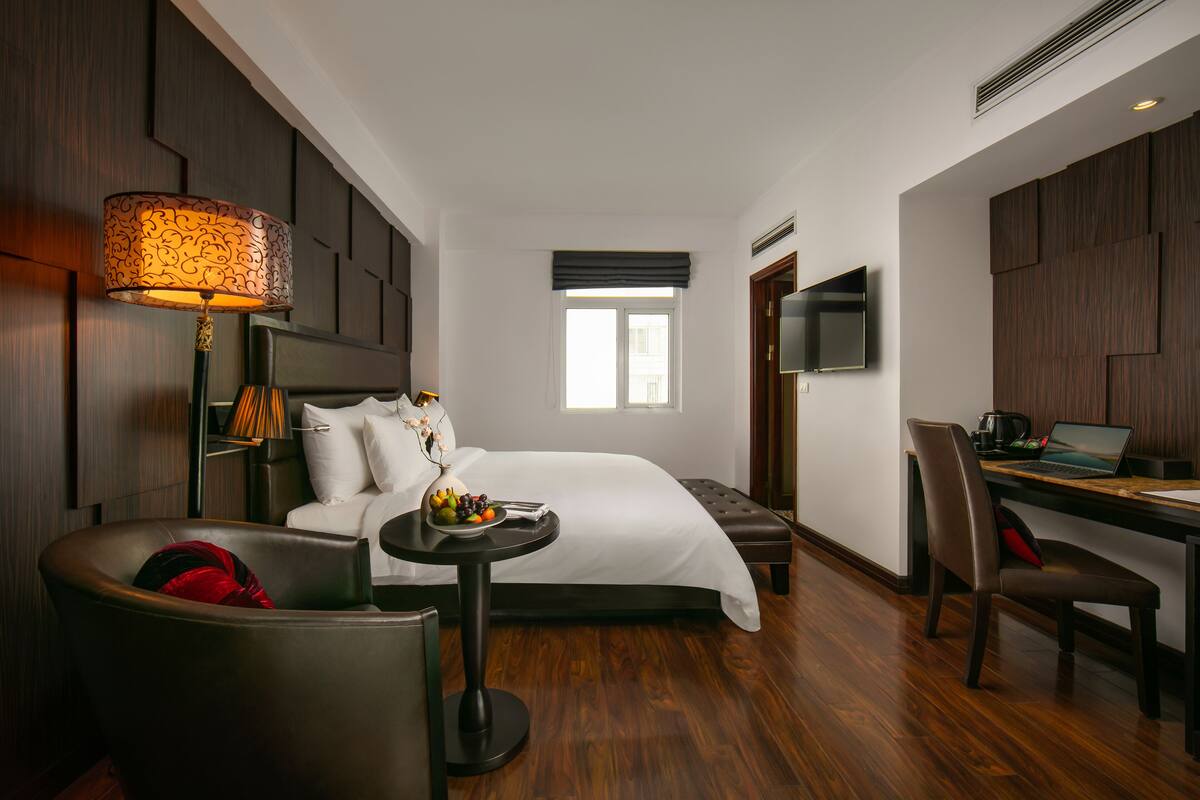 Une chambre d’hôtel avec un lit, un bureau avec un ordinateur portable, une chaise, une lampe et une petite table avec des fruits.