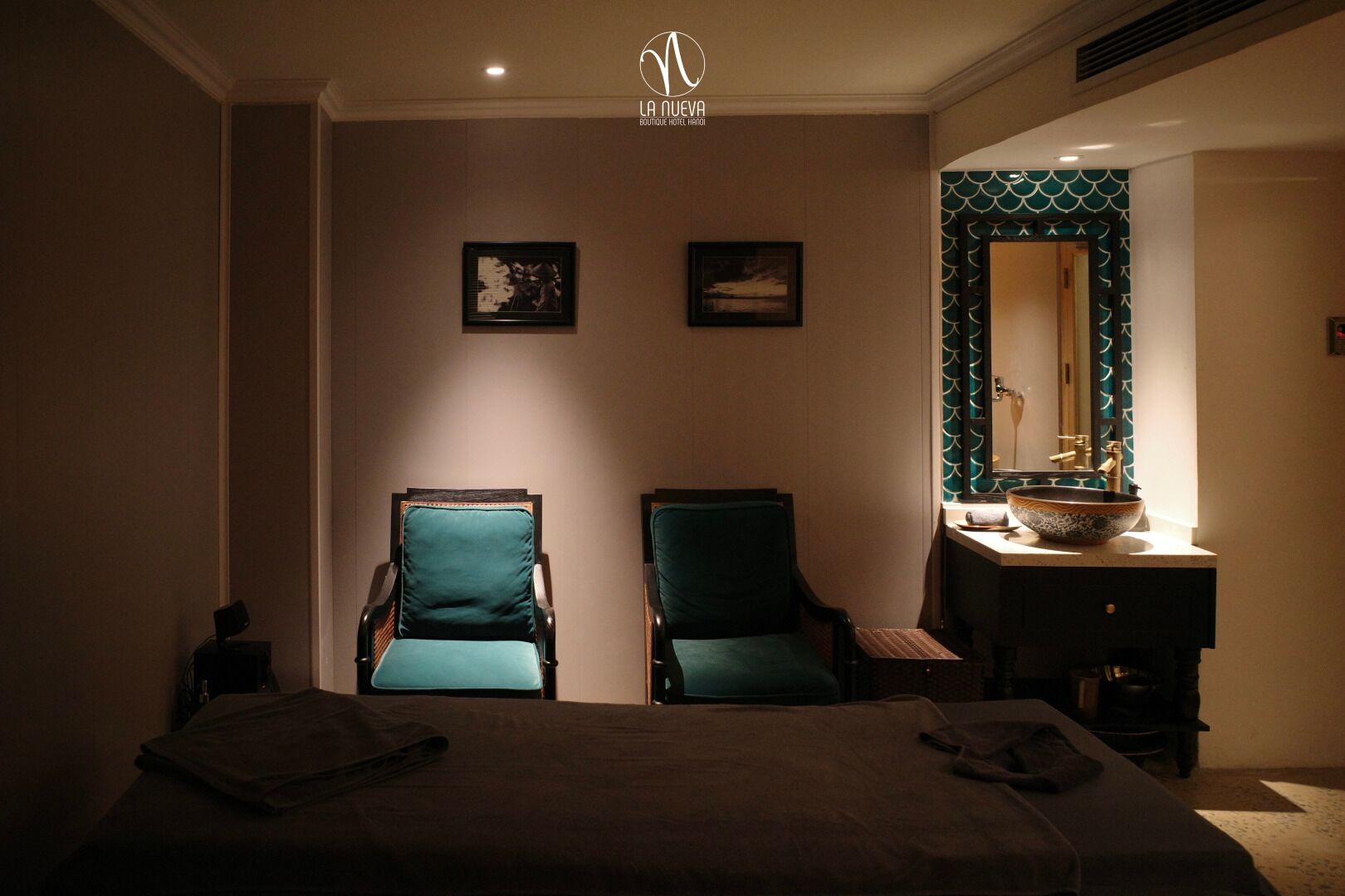 Habitación de hotel con una cama, dos sillas, un tocador con lavabo y dos cuadros en la pared.