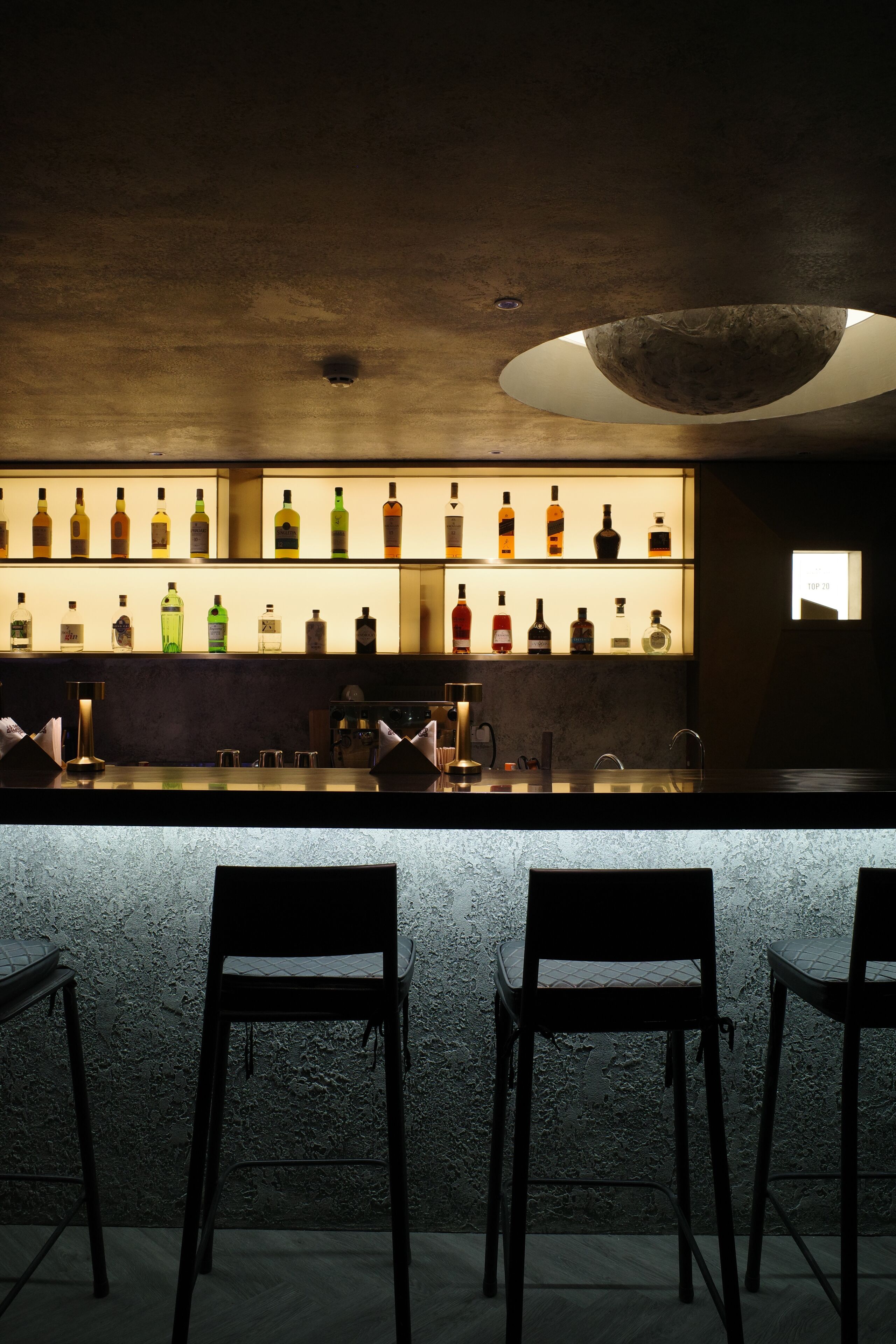 Una zona de bar con mostrador que exhibe diferentes botellas, una lámpara moderna y tres sillas.