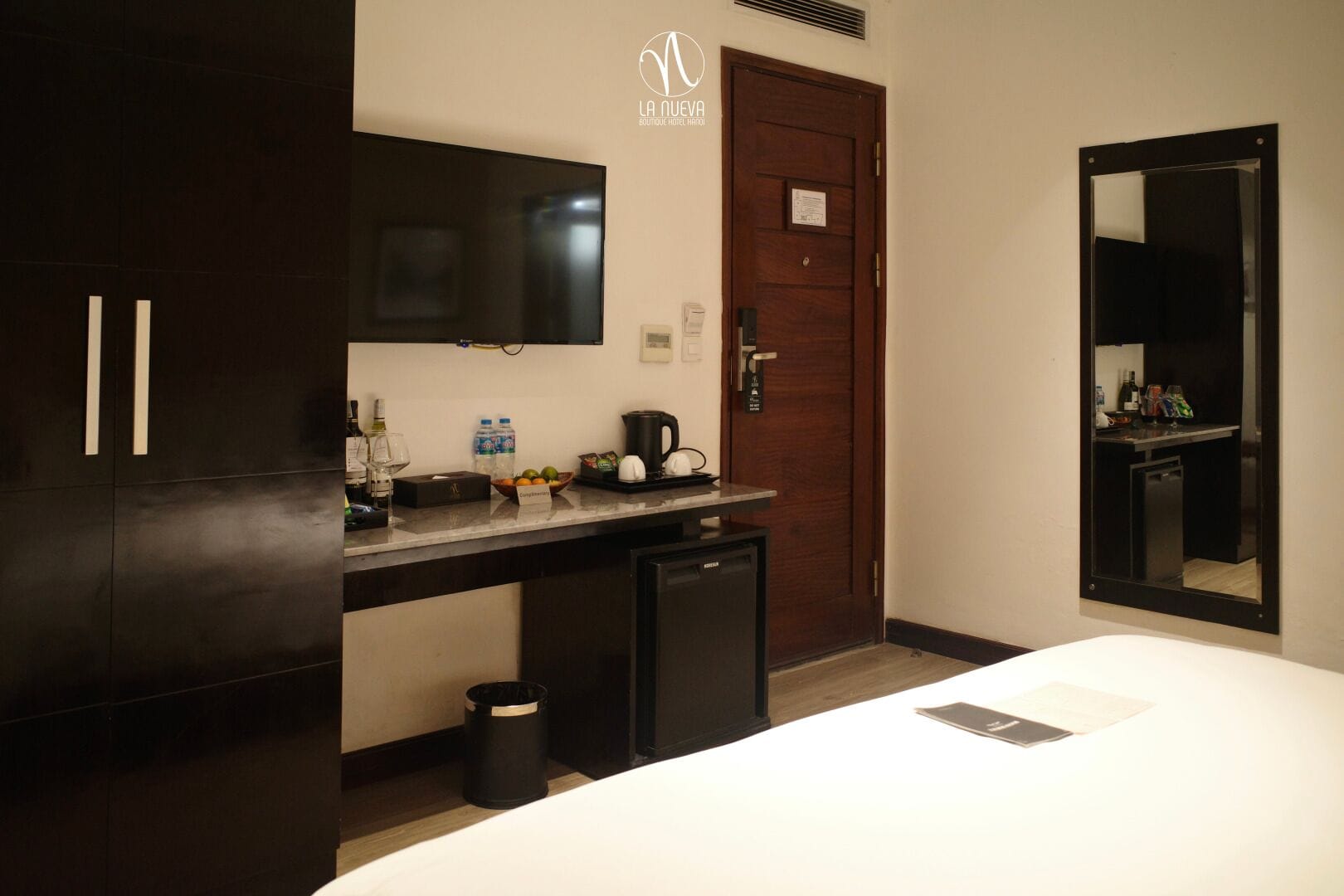 A hotel room with a bed, a small desk with a mirror, a coffee maker, and a TV mounted above the desk.