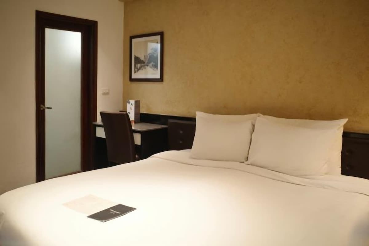 Une chambre d’hôtel avec un lit, un bureau, une chaise, un tableau encadré au mur et une porte.