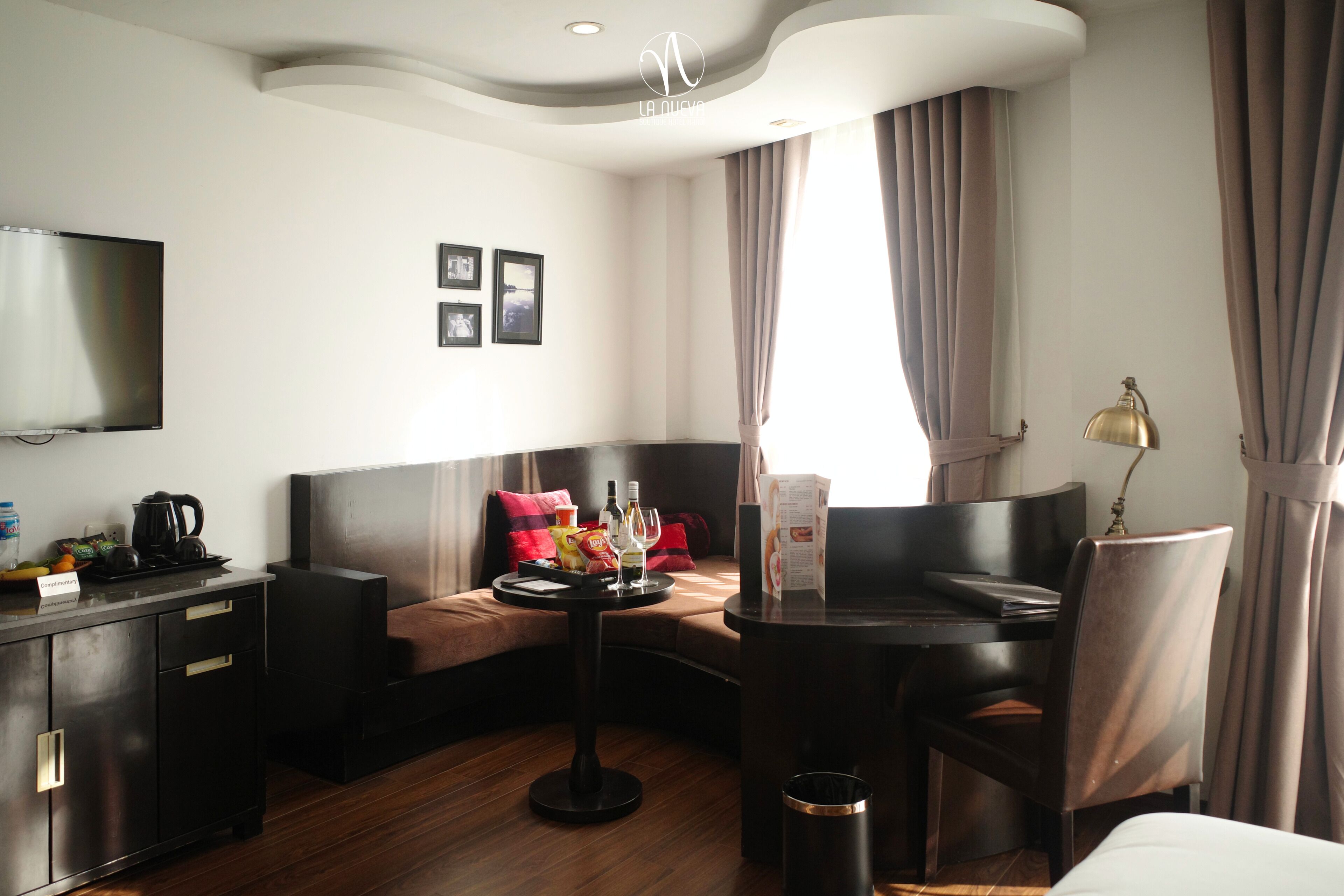 A modern hotel room with a sofa, a small round table with drinks, a TV, and a desk with a lamp.