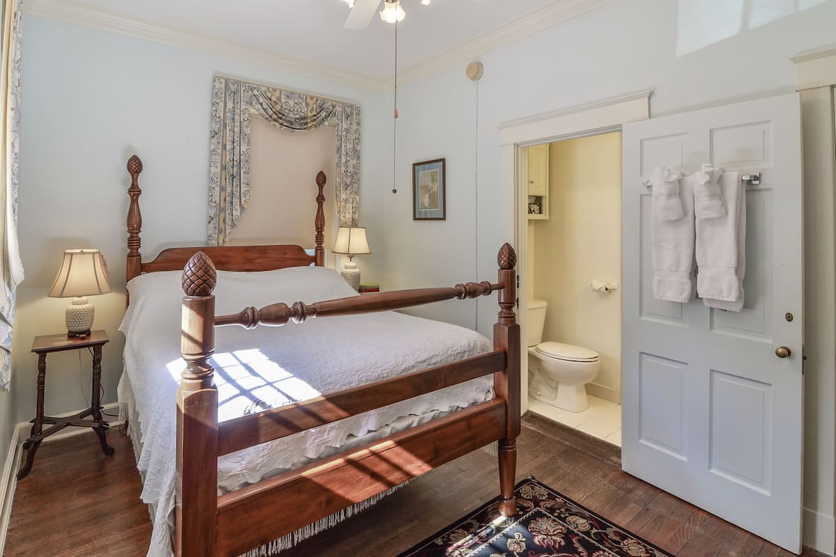 Une chambre à coucher avec un lit à baldaquin en bois, une table de chevet avec une lampe, une tapisserie à motifs et une vue sur la salle de bain, où l'on aperçoit des toilettes et des serviettes accrochées à la porte.