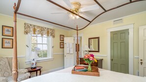 Un dormitorio con cama con dosel, ventilador de techo, ventana con cortinas florales, mesita de noche con lámpara y una puerta verde.