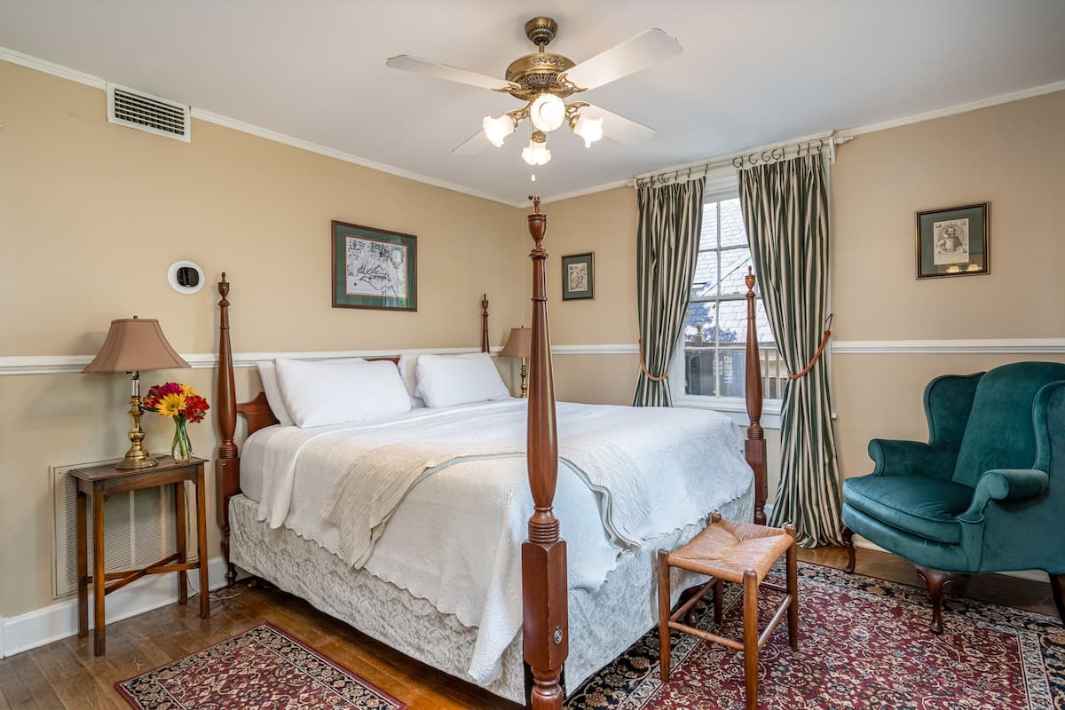 Une chambre à coucher avec un lit à baldaquin, un ventilateur de plafond, un fauteuil vert, une chaise en bois, une table de chevet avec une lampe et un vase décoré de motifs floraux.