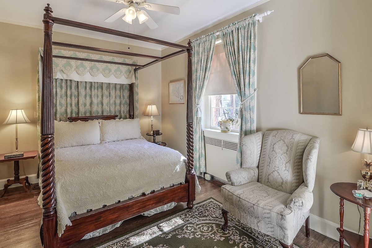 Un lit à baldaquin avec un cadre en bois et un couvre-lit blanc, une chaise assortie, une petite table avec une lampe et un grand miroir au mur.