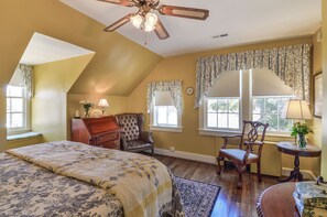 A bedroom with a large bed, a ceiling fan, a desk with a chair, and two windows with curtains.