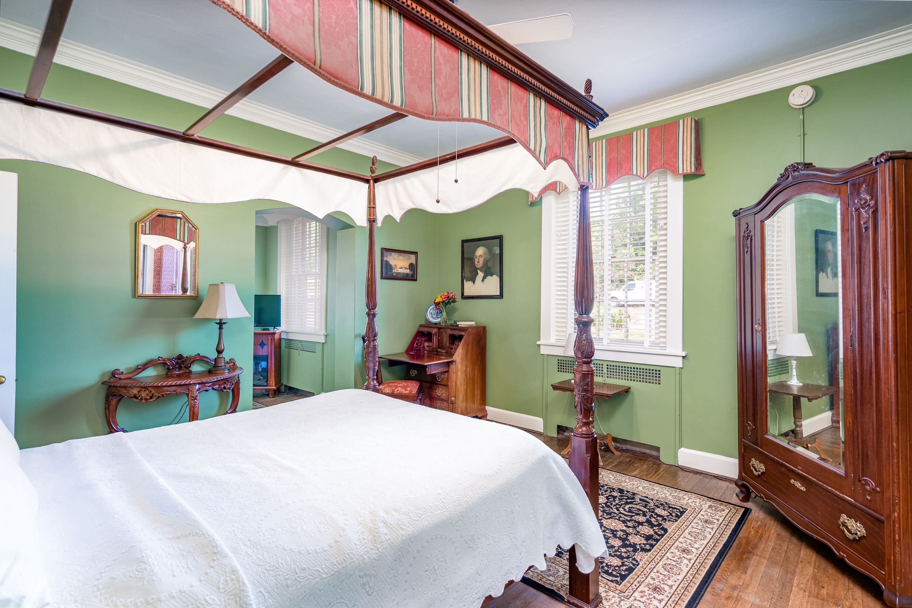 A room with a four-poster bed, a wooden wardrobe, a mirror, and a small table with a lamp.