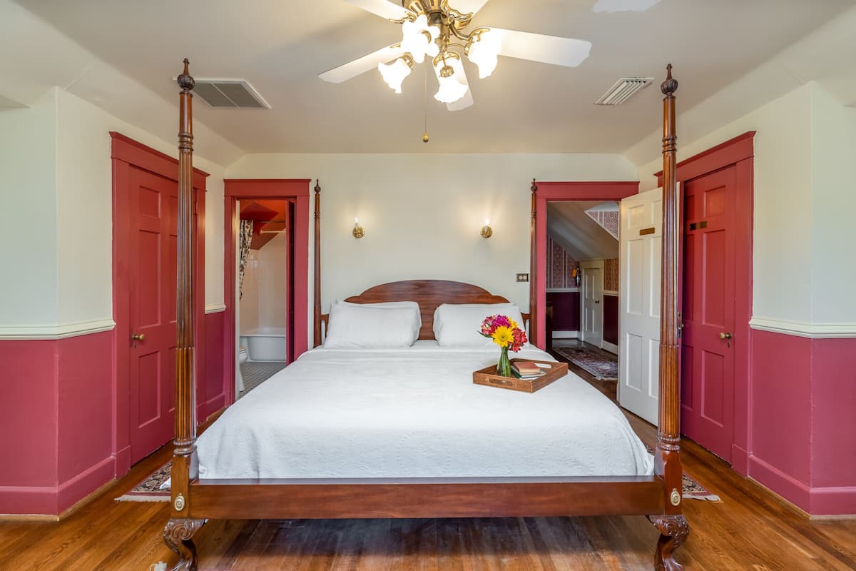Un lit à baldaquin avec une tête de lit en bois, un ventilateur de plafond et une table de chevet sur laquelle est posé un plateau de fleurs.