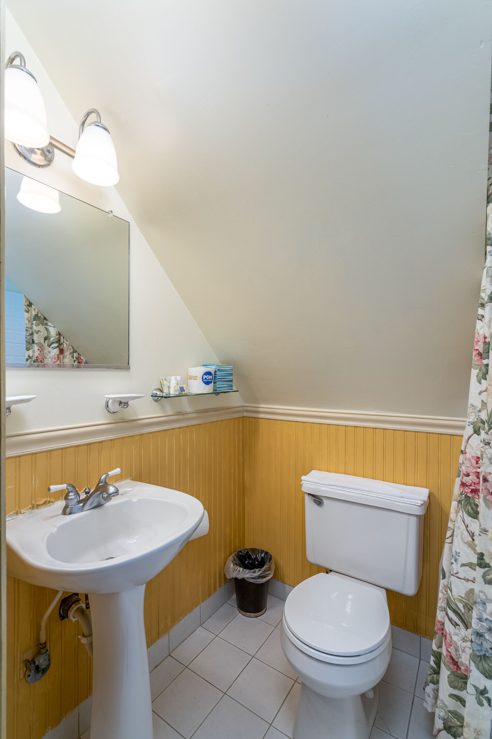 A bathroom with a white sink, a toilet, and a mirror. There is a trash can and a shelf with toiletries.