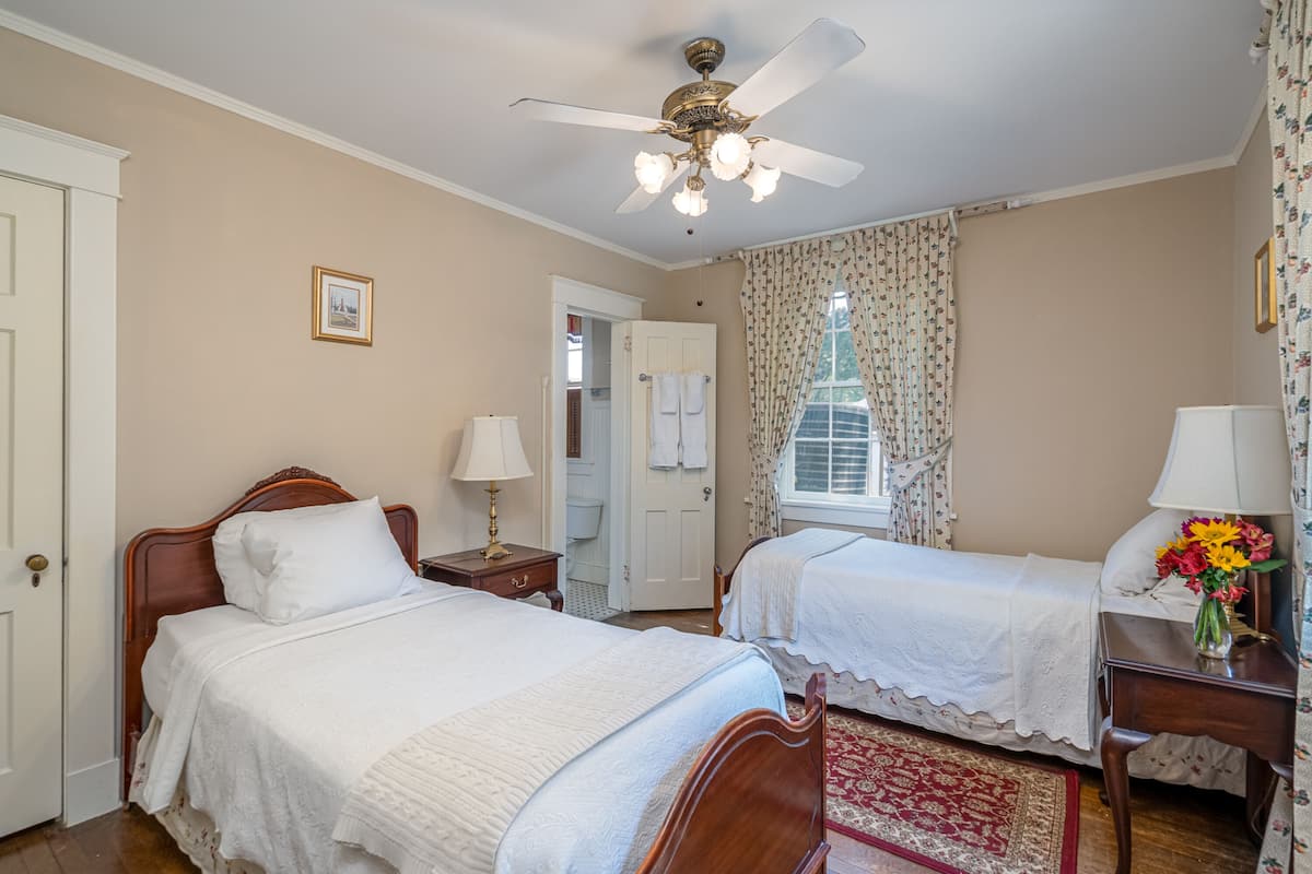 Une chambre avec deux lits, un ventilateur de plafond, une fenêtre avec des rideaux et une table de chevet avec une lampe et des fleurs.