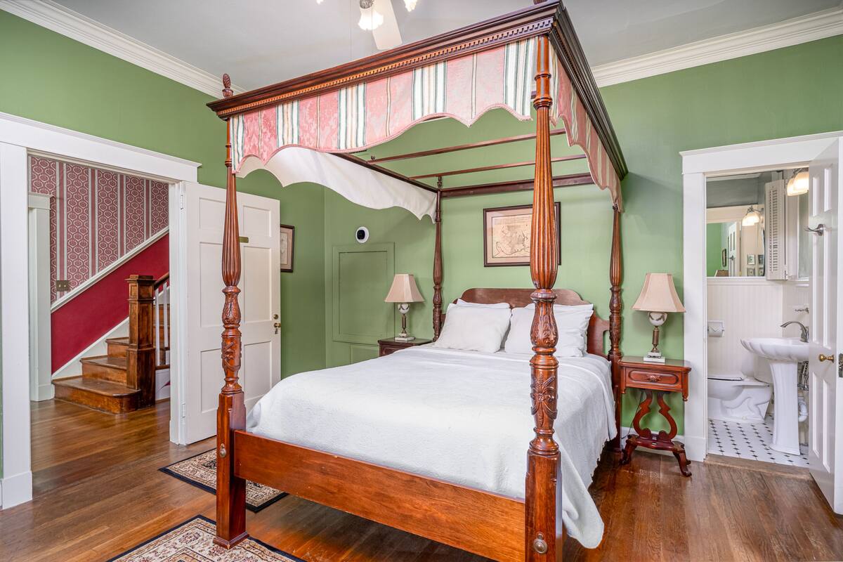 Un lit à baldaquin avec des rideaux blancs, une table de chevet en bois, une salle de bain blanche avec un lavabo et des toilettes, et un escalier avec une rampe rouge.