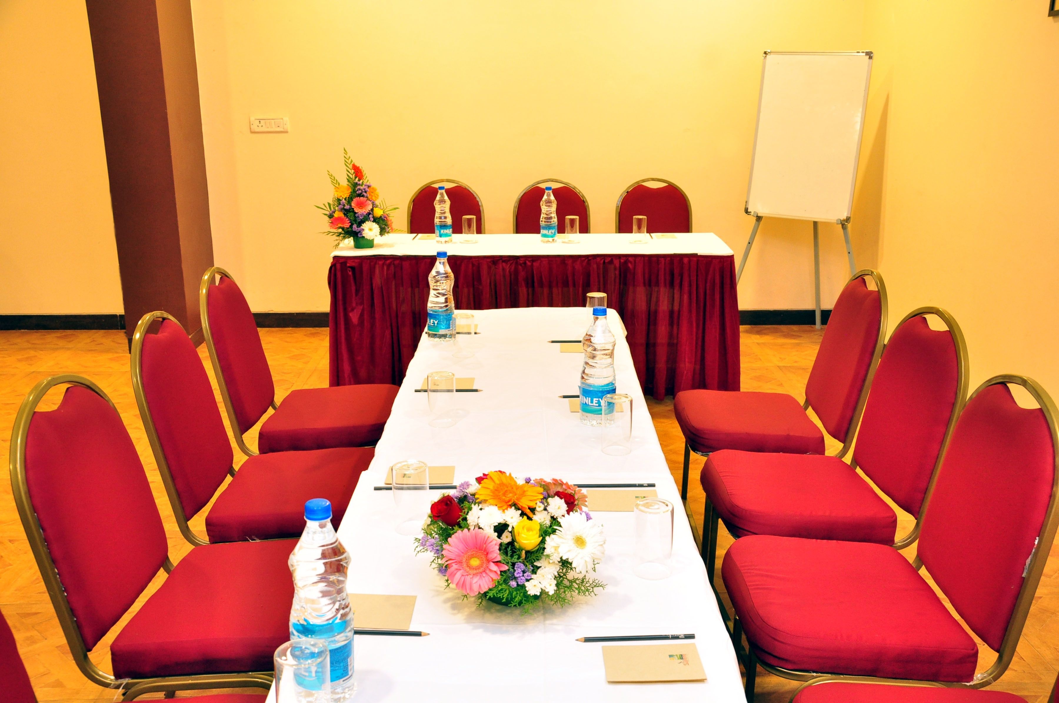 Une salle de conférence avec une longue table, des chaises rouges, des bouteilles d’eau, un arrangement floral au centre et un tableau blanc.