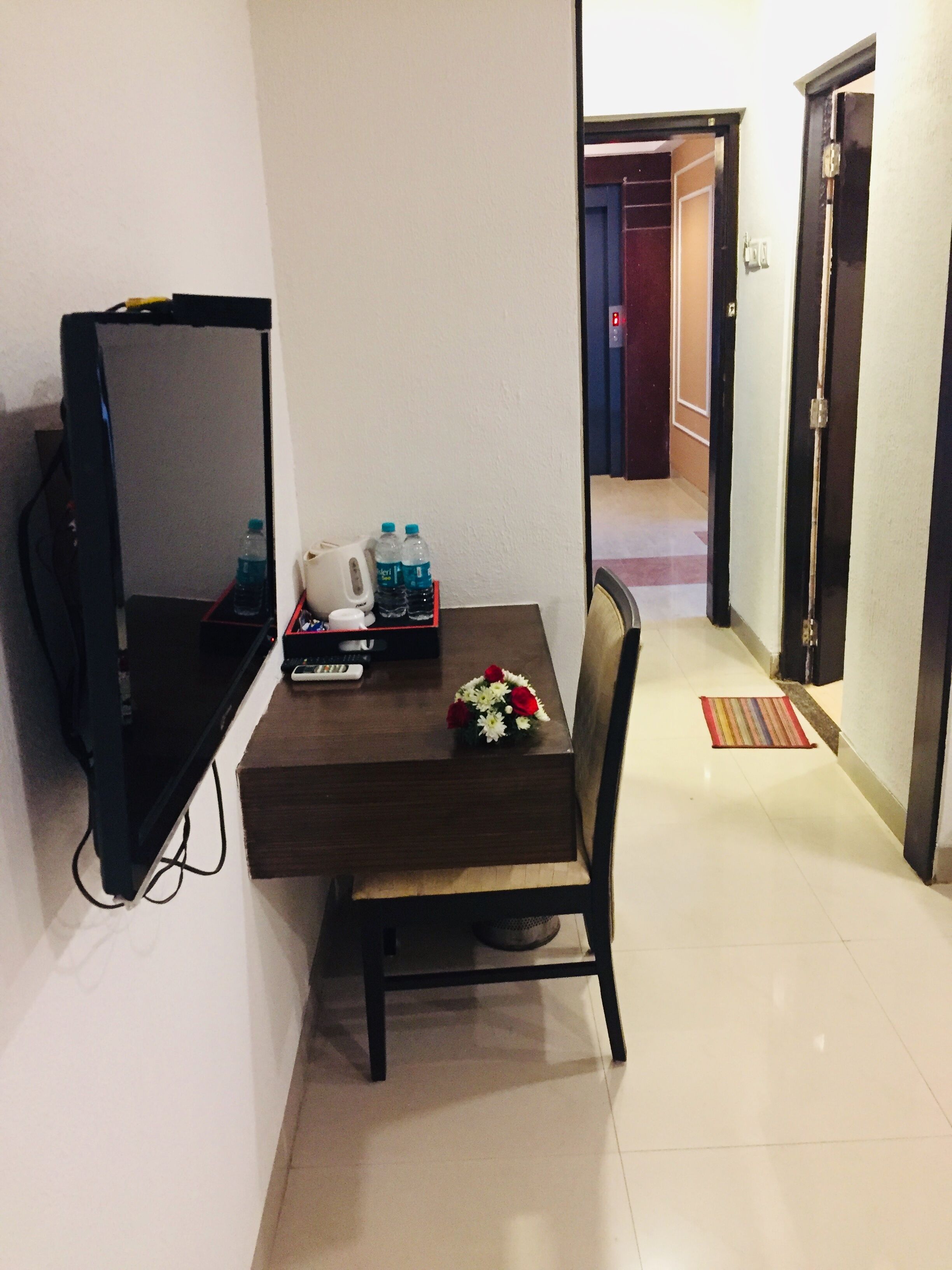 A small room with a TV on a stand, a table with a chair, a bouquet of flowers, and a water bottle.