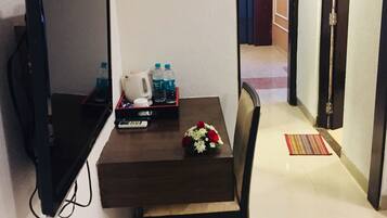 A small room with a TV on a stand, a table with a chair, a bouquet of flowers, and a water bottle.