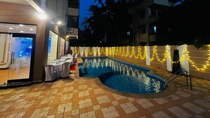 An outdoor swimming pool area with a tiled floor, a pool with water fountains, and a building entrance with a glass door. - Hotel Supreme (Vasco) (Vasco da Gama)