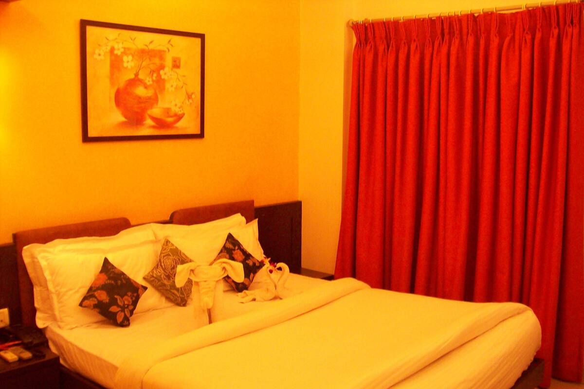 Una cama bien hecha con sábanas blancas y almohadones decorativos, una obra de arte enmarcada en la pared y una cortina roja.