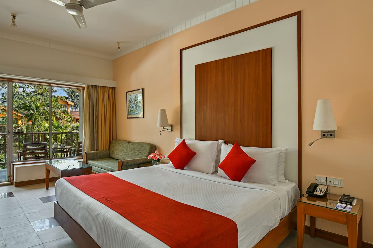 A hotel room with a large bed, a desk, a chair, a balcony with a view of palm trees, and a ceiling fan.