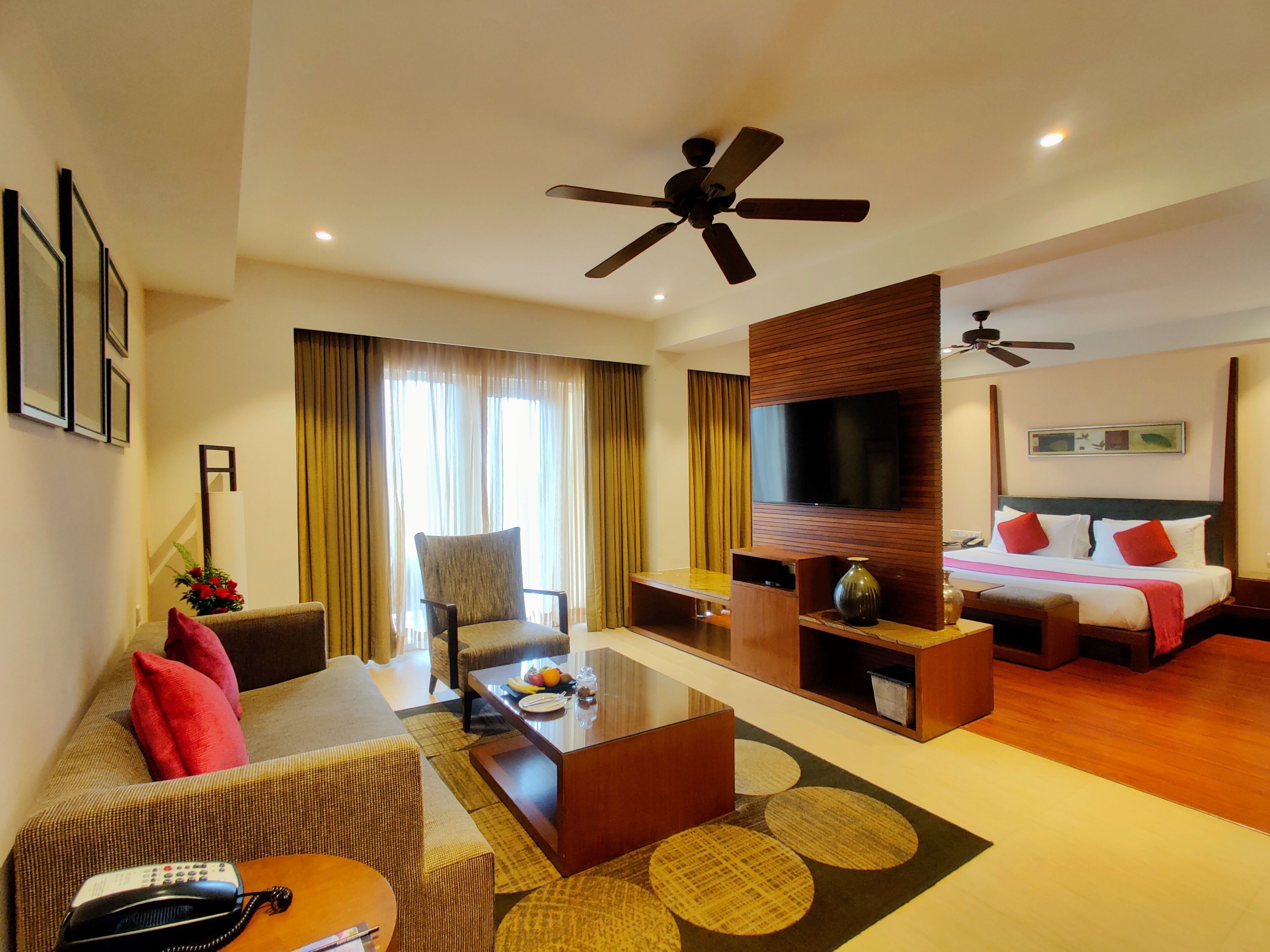 A hotel room with a sofa, armchair, coffee table, TV, and a bed with red pillows.