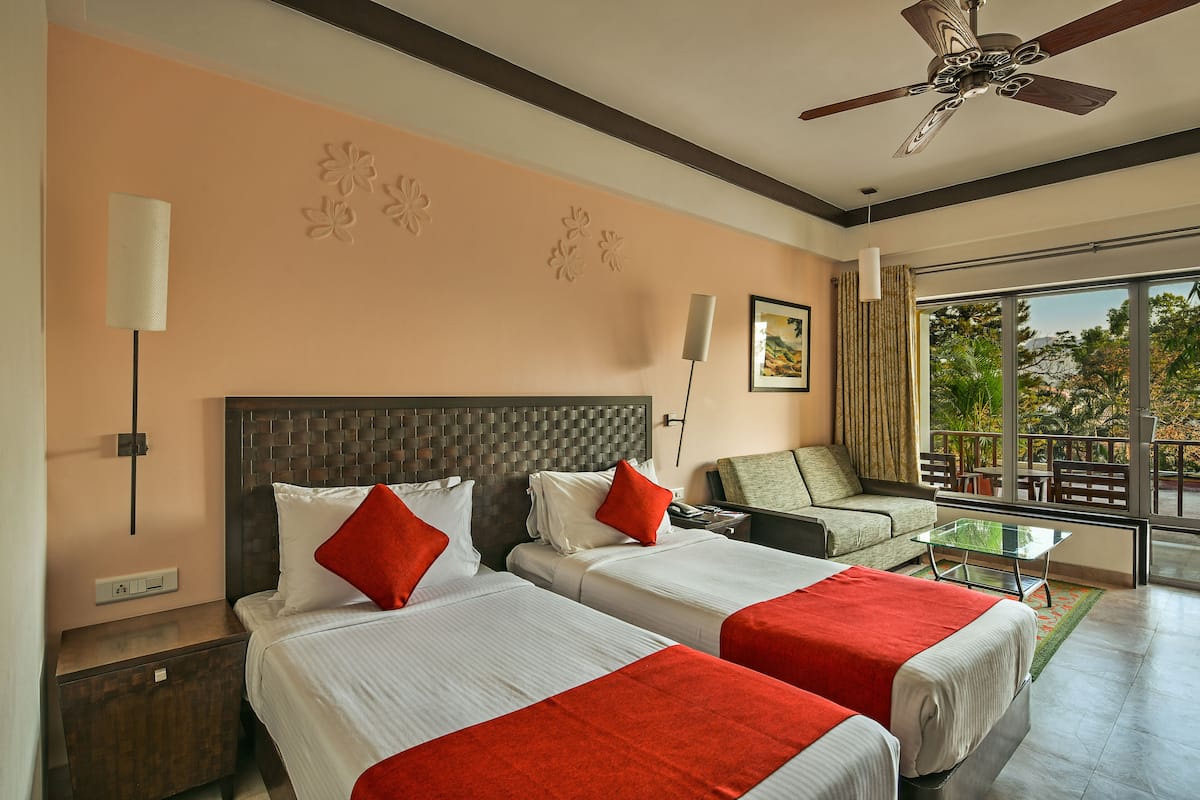 A hotel room with two beds, a ceiling fan, a large window with a view of greenery, and a balcony with outdoor furniture.