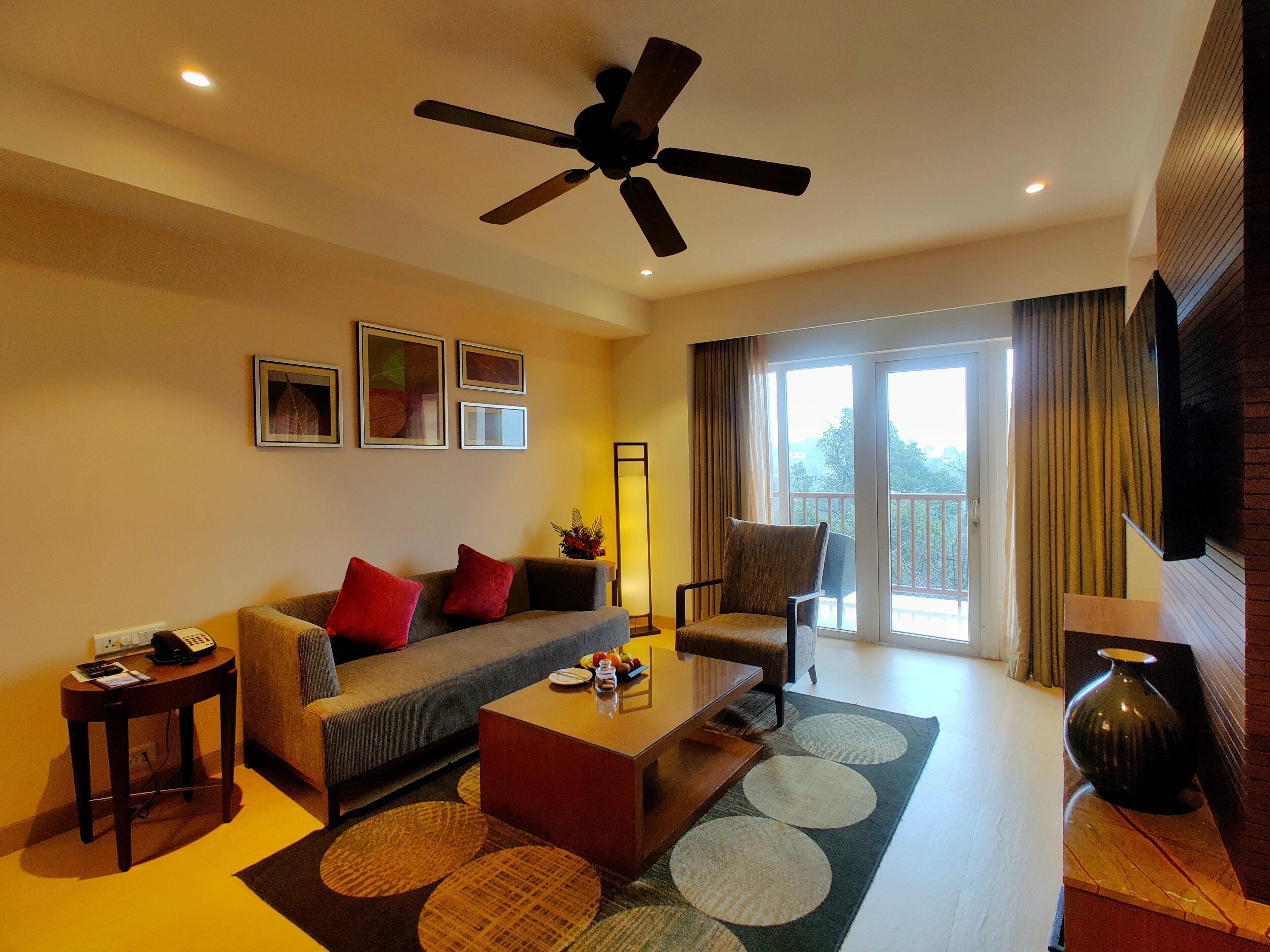 A living room with a sofa, coffee table, and a ceiling fan. There are framed pictures on the wall and a TV mounted on the wall.