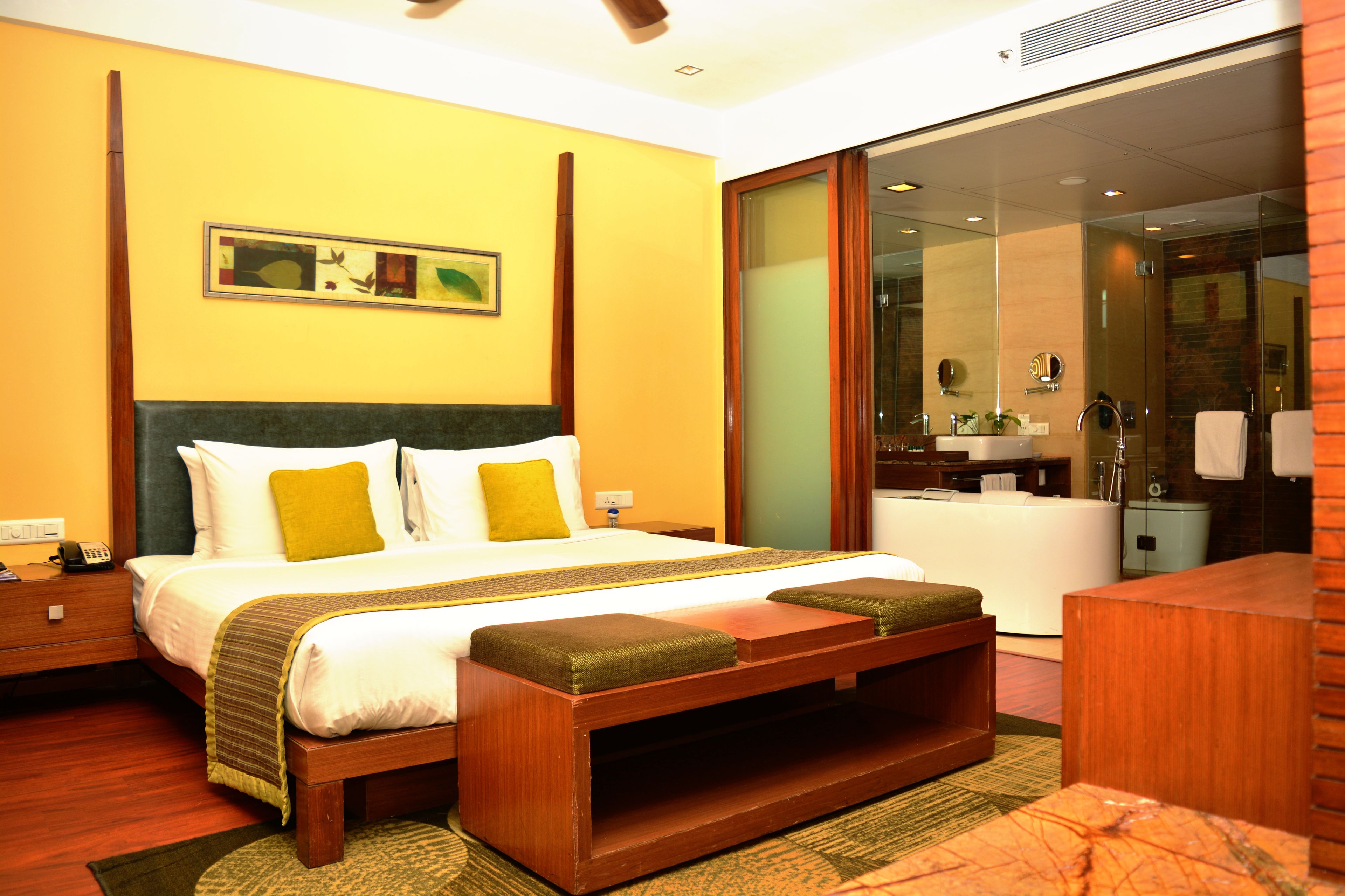 A hotel room with a large bed, a wooden bench, a bathroom with a glass shower, and a painting on the wall.