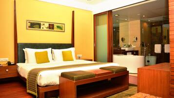 A hotel room with a large bed, a wooden bench, a bathroom with a glass shower, and a painting on the wall.