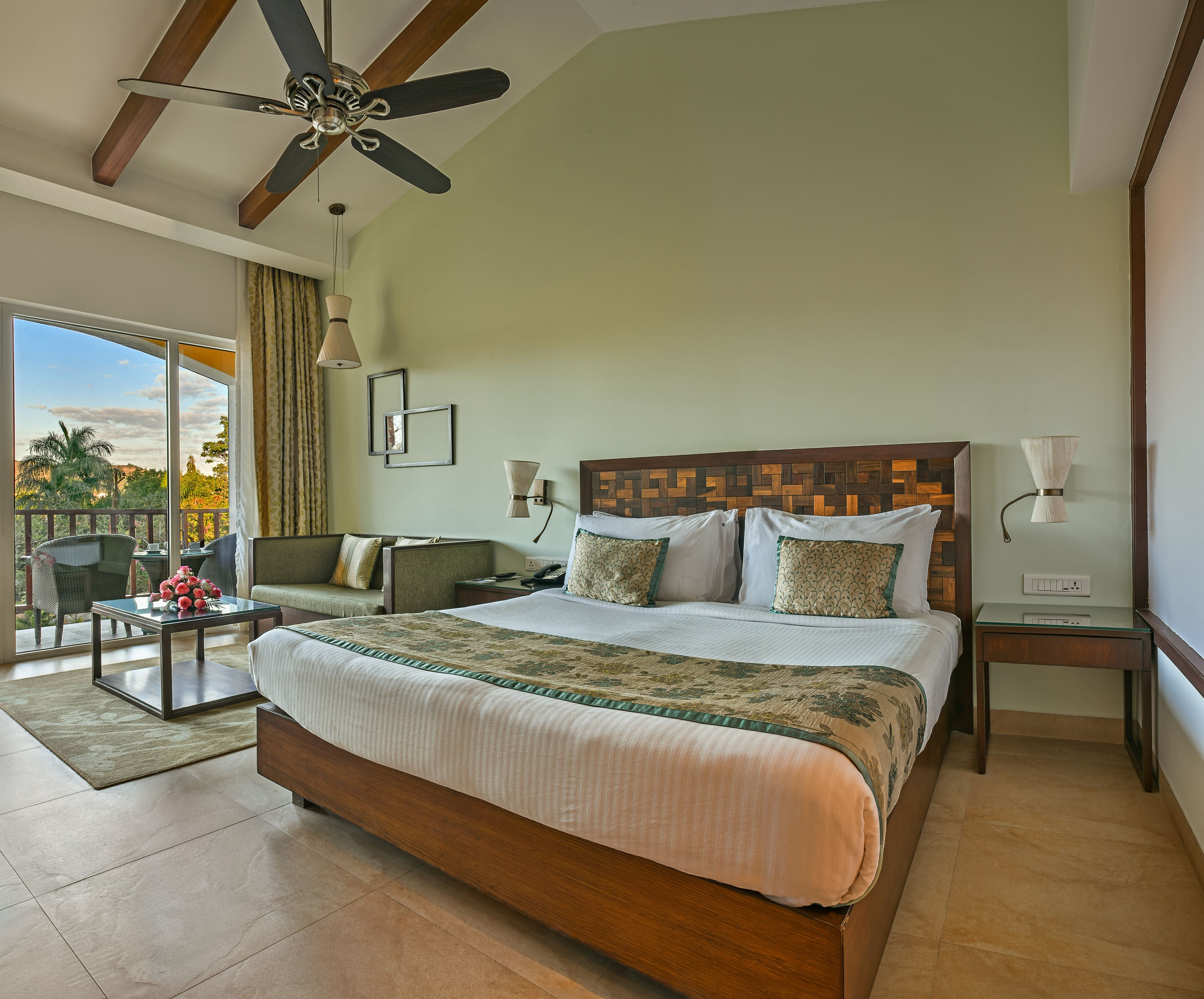 A spacious bedroom with a large bed, a ceiling fan, a balcony with a view, and a wooden headboard.