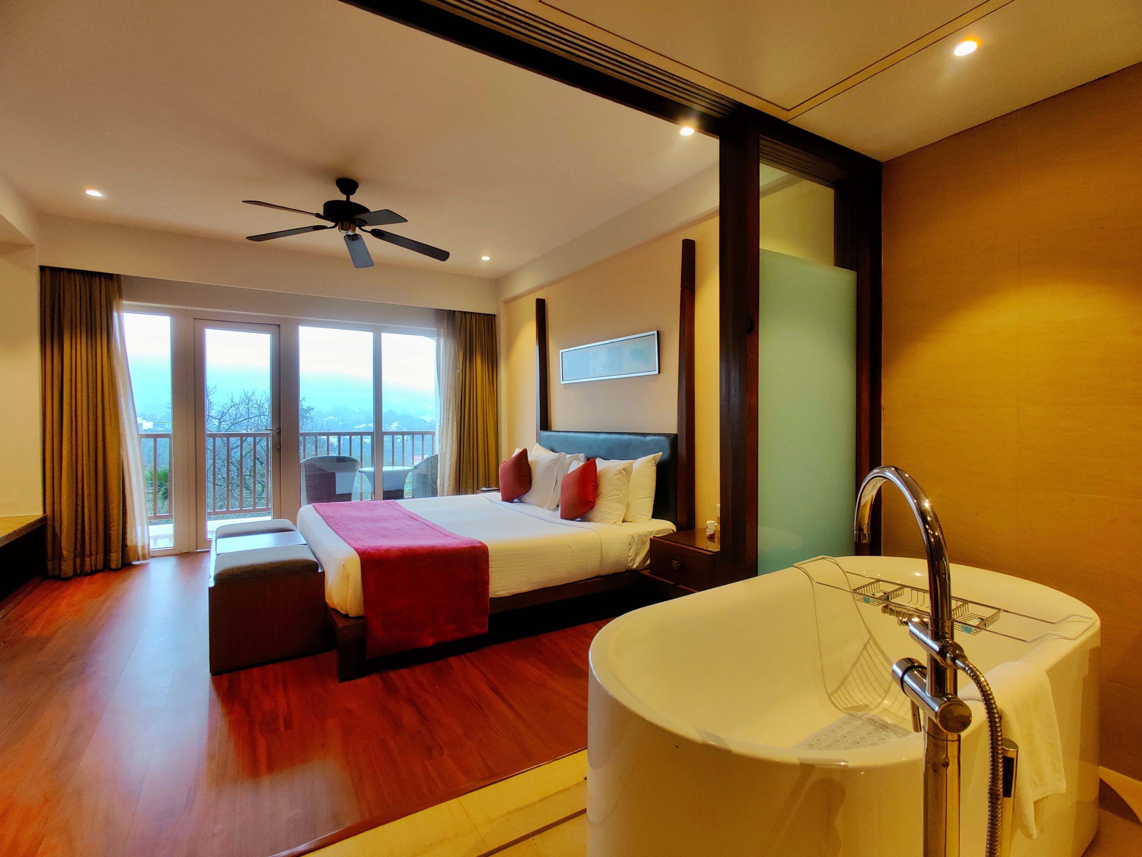 A hotel room with a bed, a large bathtub, a ceiling fan, and a balcony with a view.
