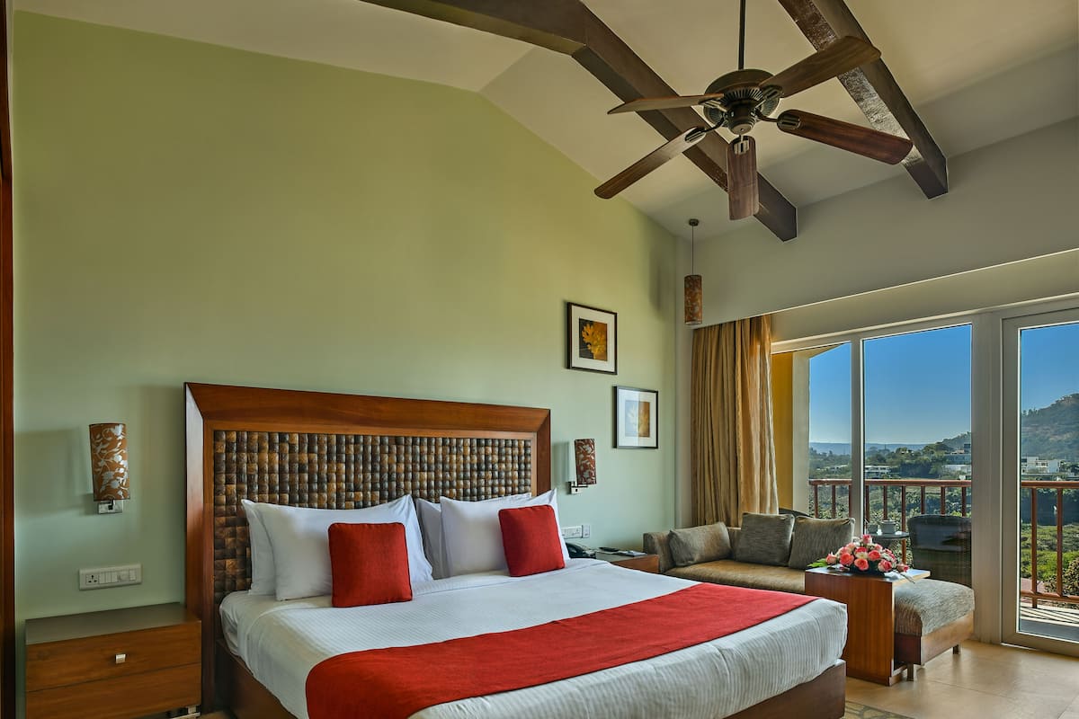 A spacious bedroom with a large bed, a ceiling fan, a balcony with a view, and a wooden headboard.