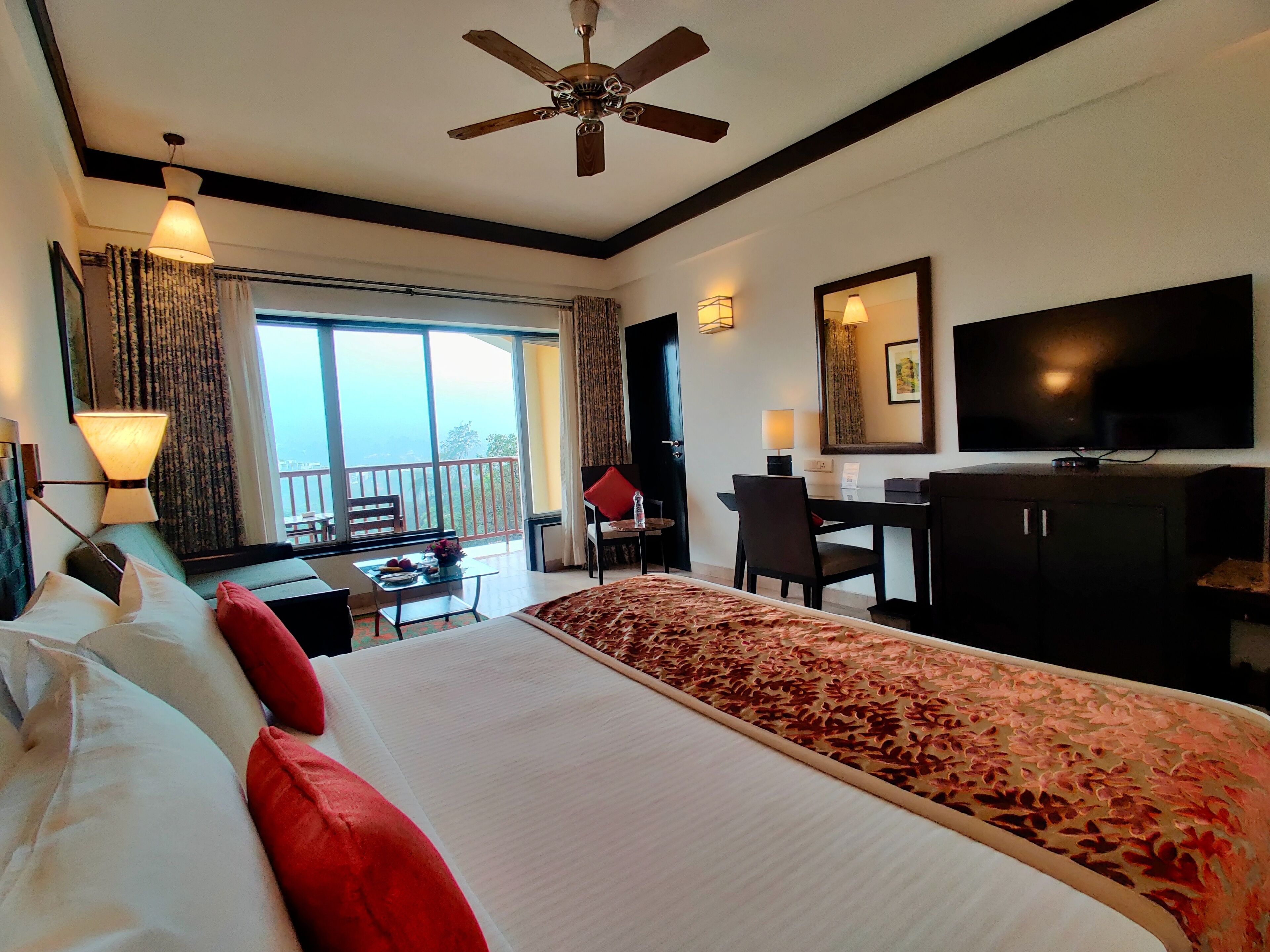 A hotel room with a large bed, a desk, a chair, a television, and a balcony with curtains.
