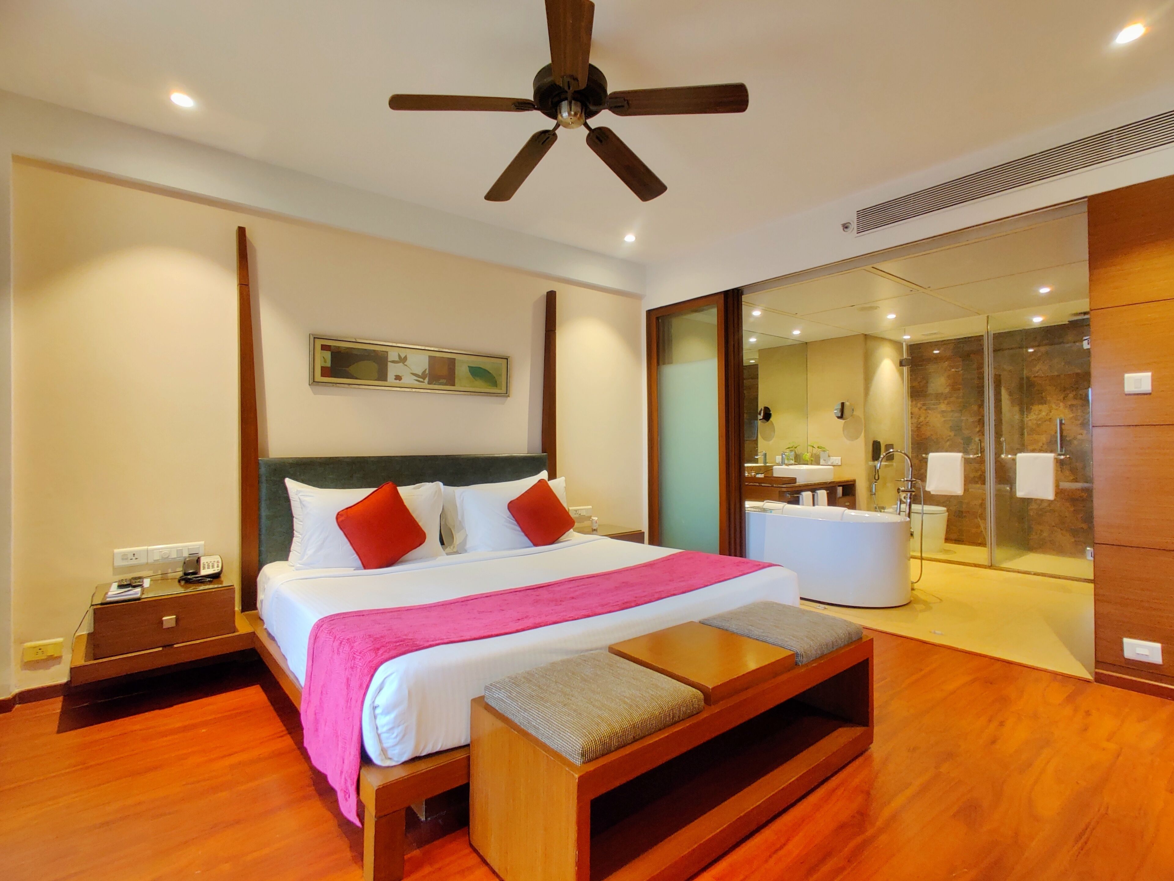 A hotel room with a bed, bedside table, and a small wooden bench. There is a ceiling fan, a bathroom with a glass shower, and a mirror.