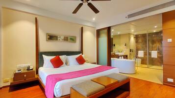 A hotel room with a bed, bedside table, and a small wooden bench. There is a ceiling fan, a bathroom with a glass shower, and a mirror.