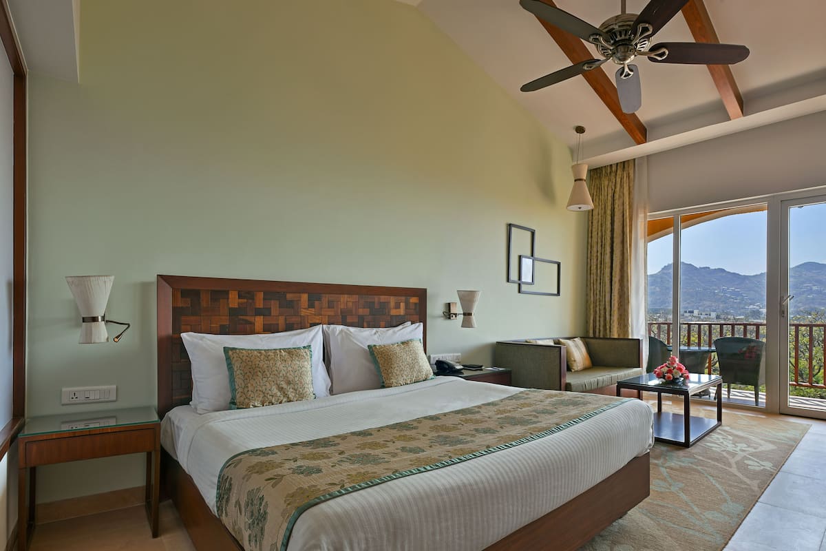A hotel room with a large bed, a desk, a chair, a balcony with a view, and a ceiling fan.