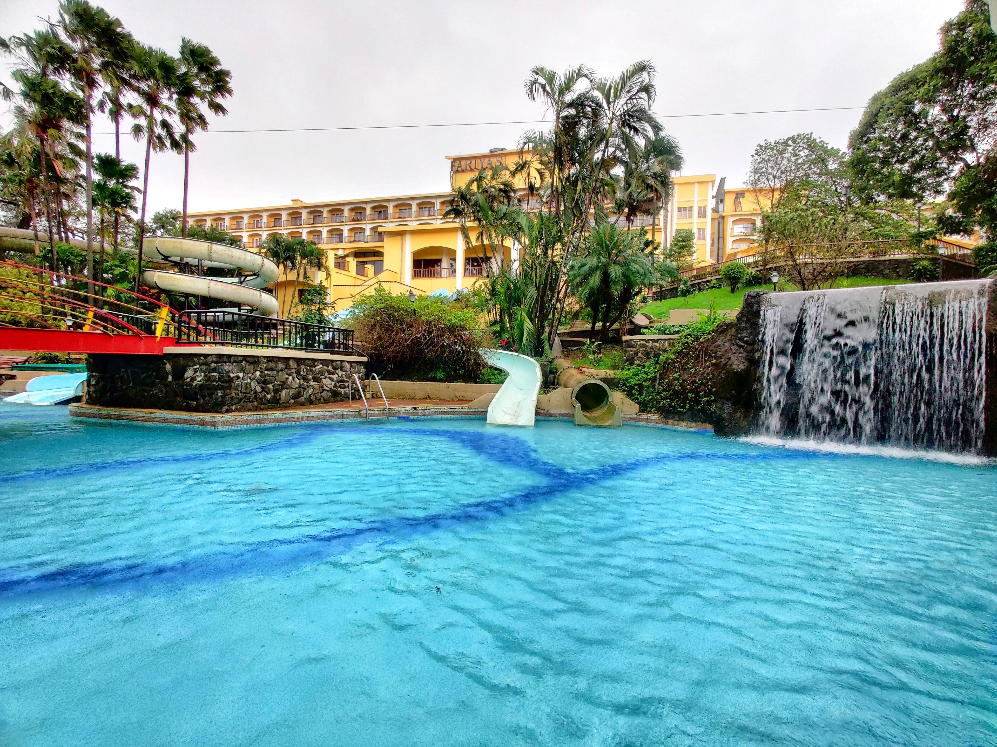 A swimming pool with a waterfall feature, a red bridge, and a yellow building in the background.