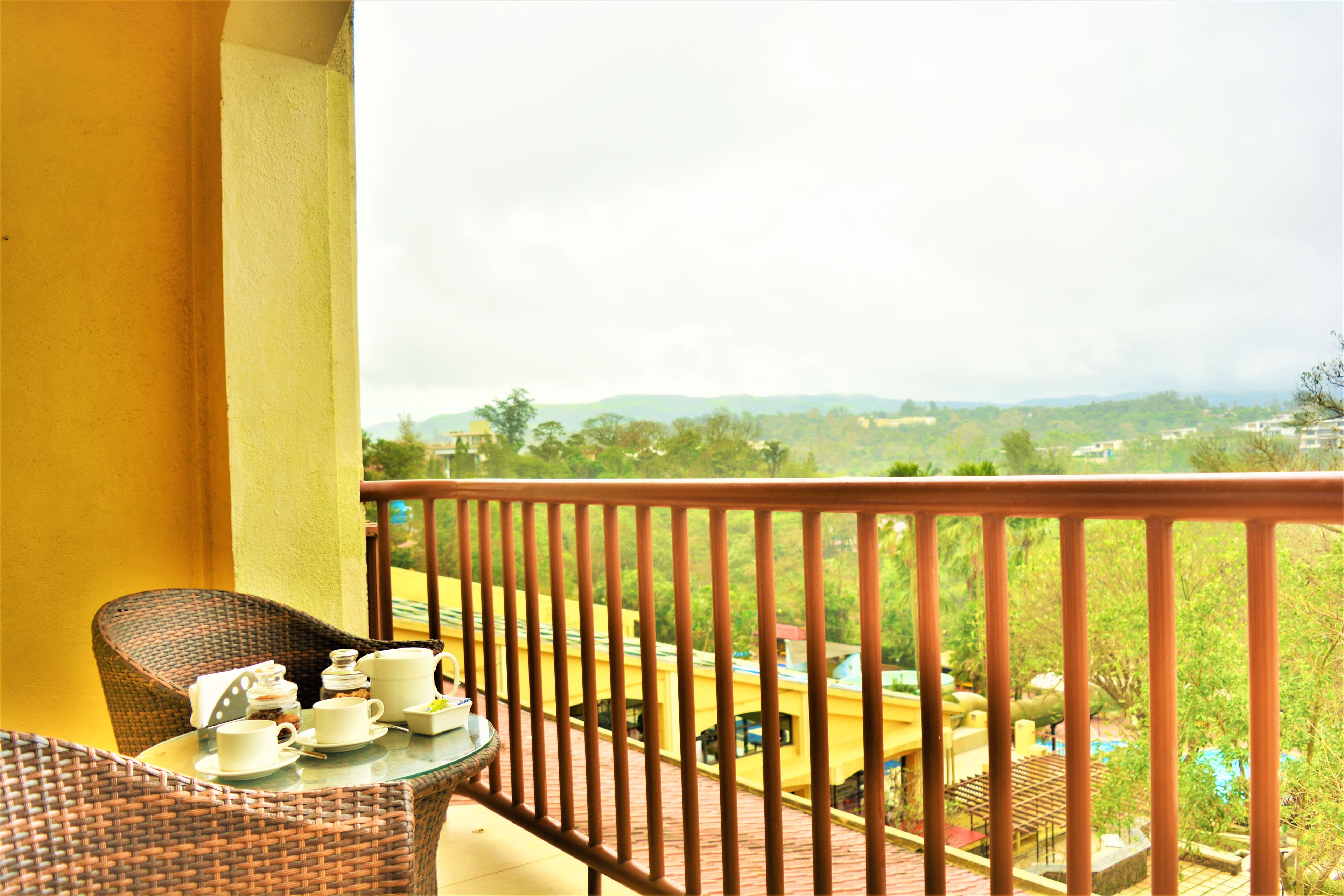 A balcony with wicker furniture, a table with cups and saucers, and a view of a landscape with buildings and trees.