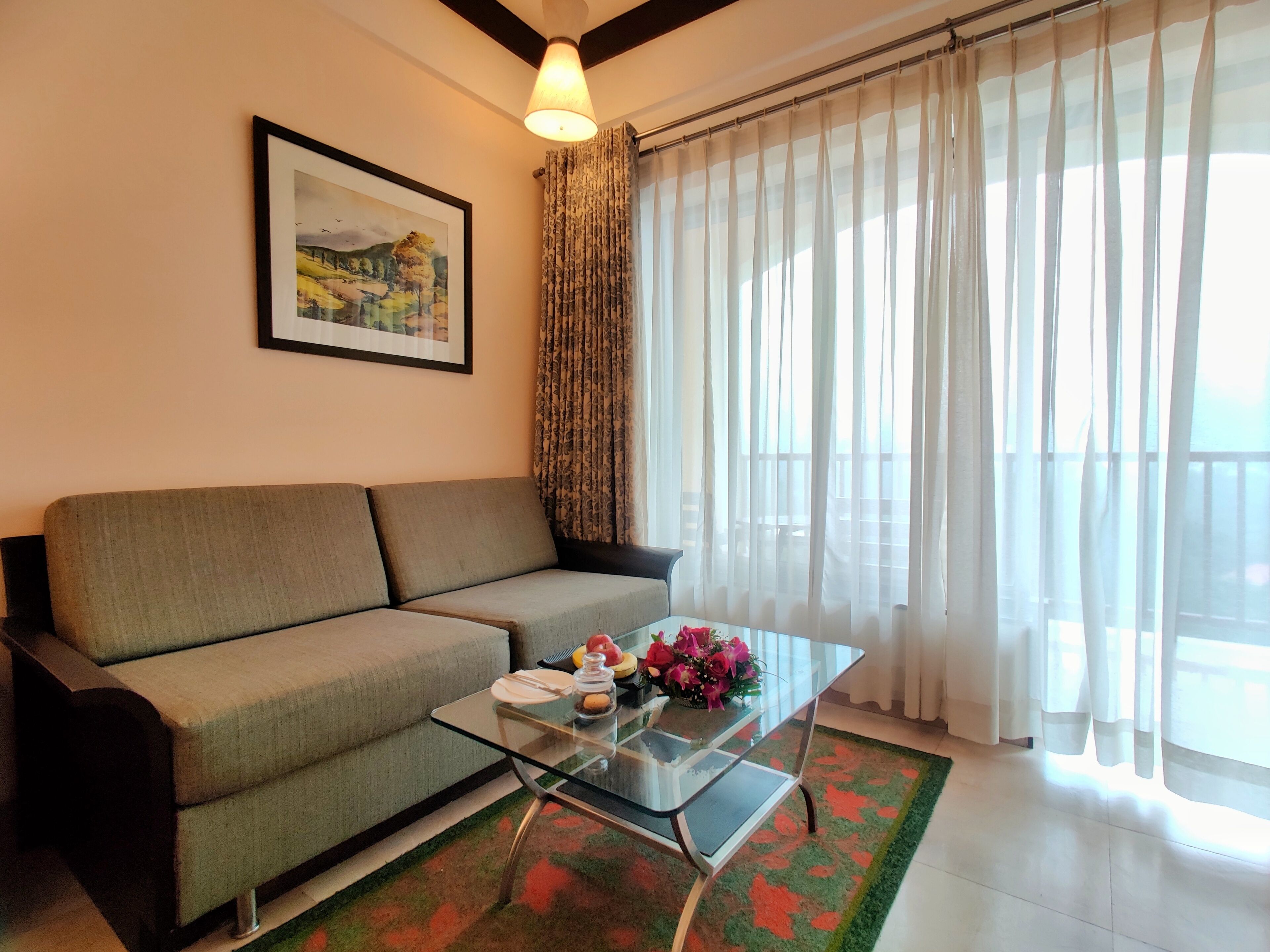 A living room with a sofa, a glass coffee table, a framed painting, and sheer curtains.