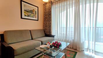 A living room with a sofa, a glass coffee table, a framed painting, and sheer curtains.