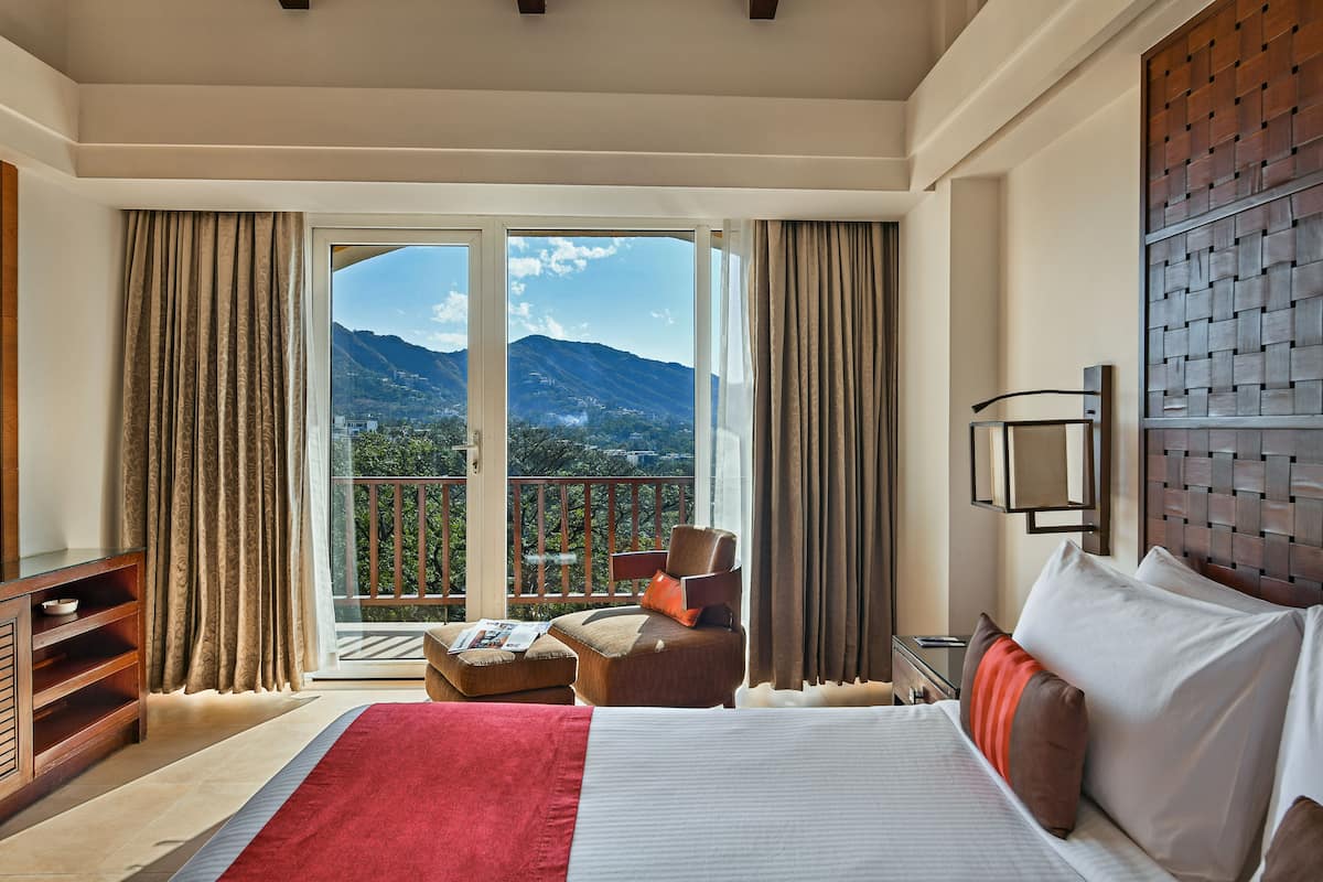 A hotel room with a large bed, a chair, a small table with a magazine, and a balcony with a mountain view.