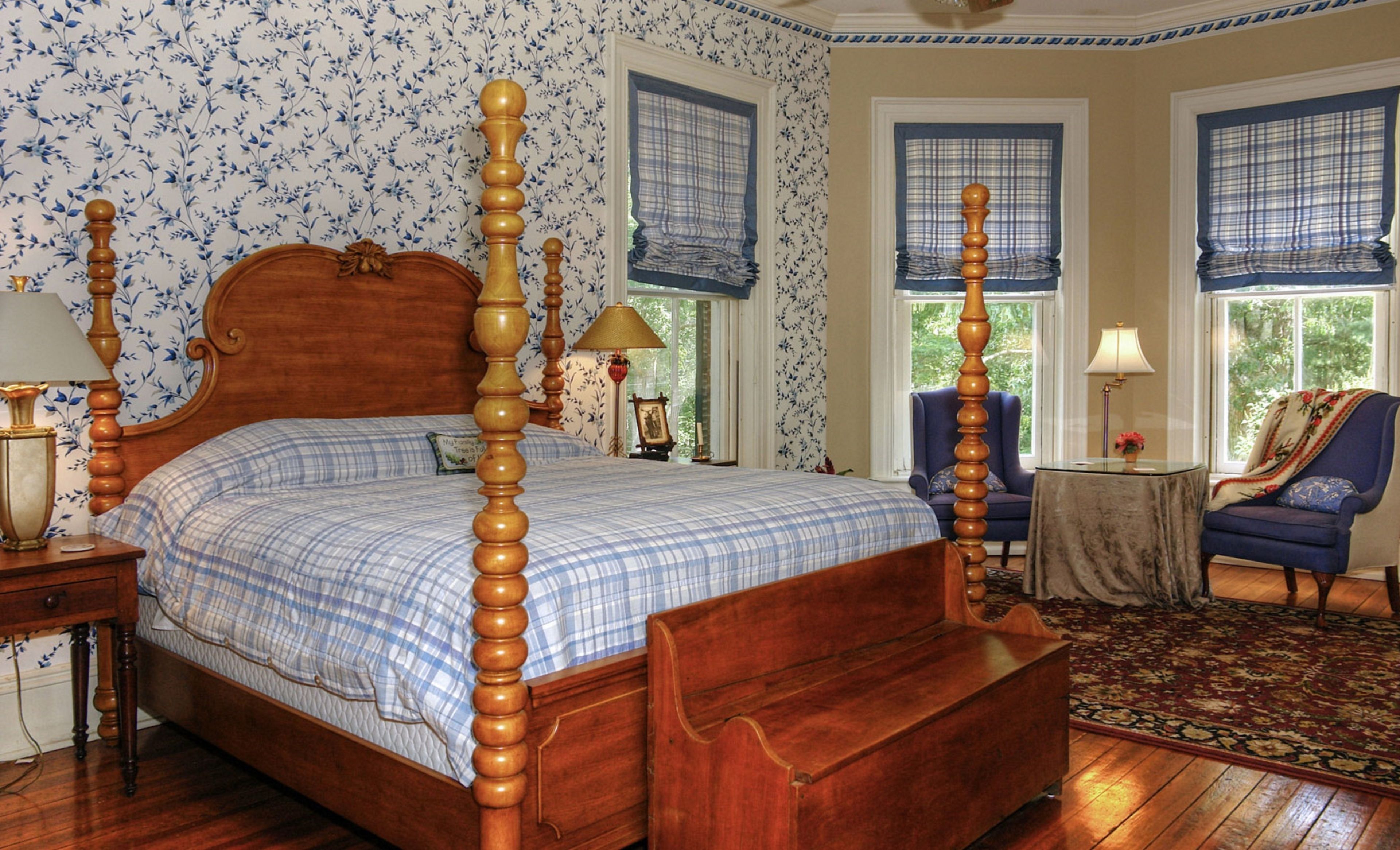 A four-poster bed with a wooden headboard and footboard, flanked by two nightstands with lamps. The room features floral wallpaper, a blue plaid bedspread, and a patterned rug on a wooden floor.