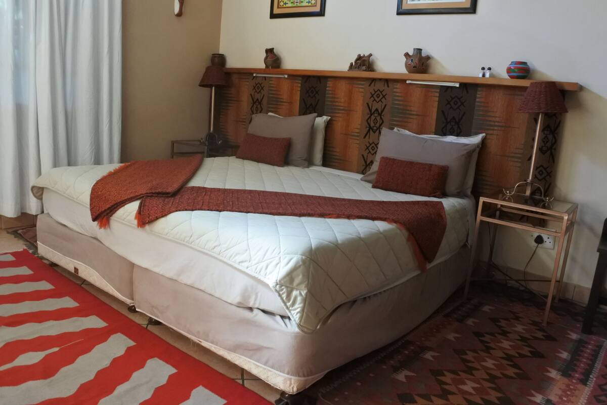 Een bed met een gewatteerd matras, een rode plaid en een hoofdbord met een gedessineerd patroon. Er staat een nachtkastje met een lamp en een klein plankje met decoratieve items.
