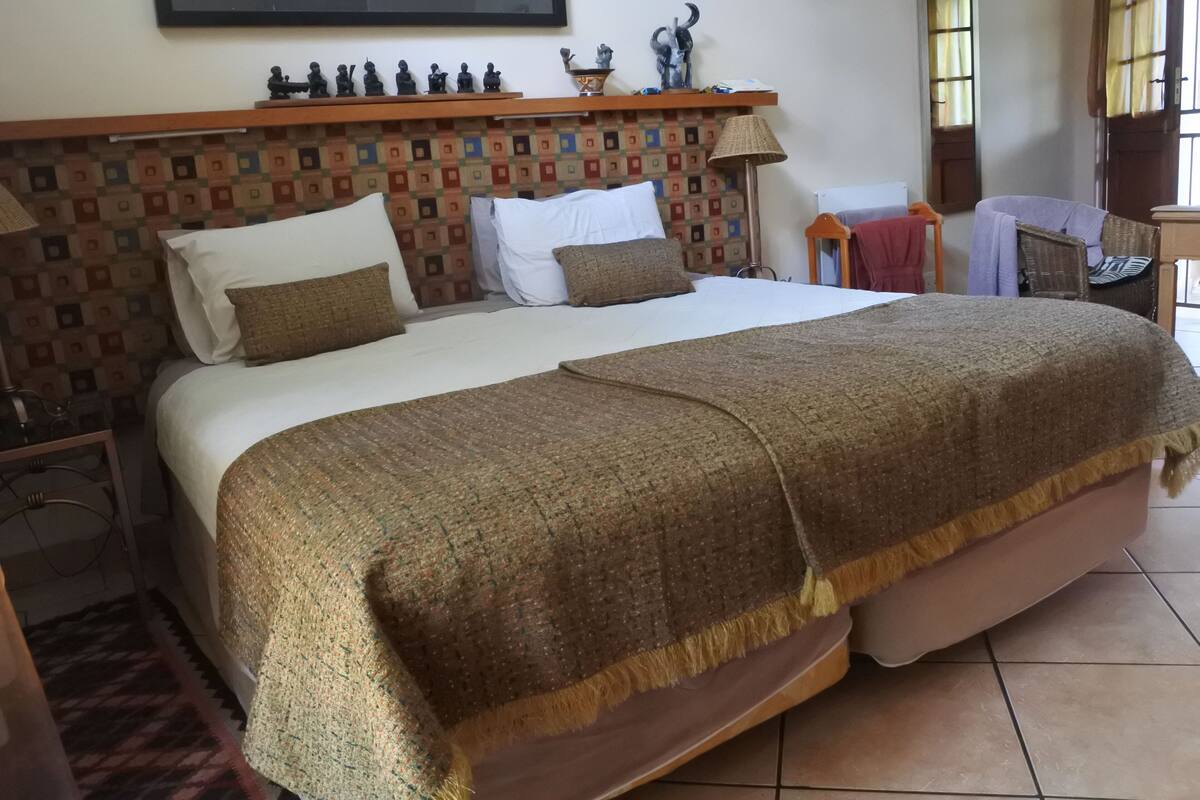 Een netjes opgemaakt bed met een bedovertrek en kussens, een hoofdbord met een geometrisch ontwerp, een klein tafeltje met een lamp, en een raam met gordijnen.