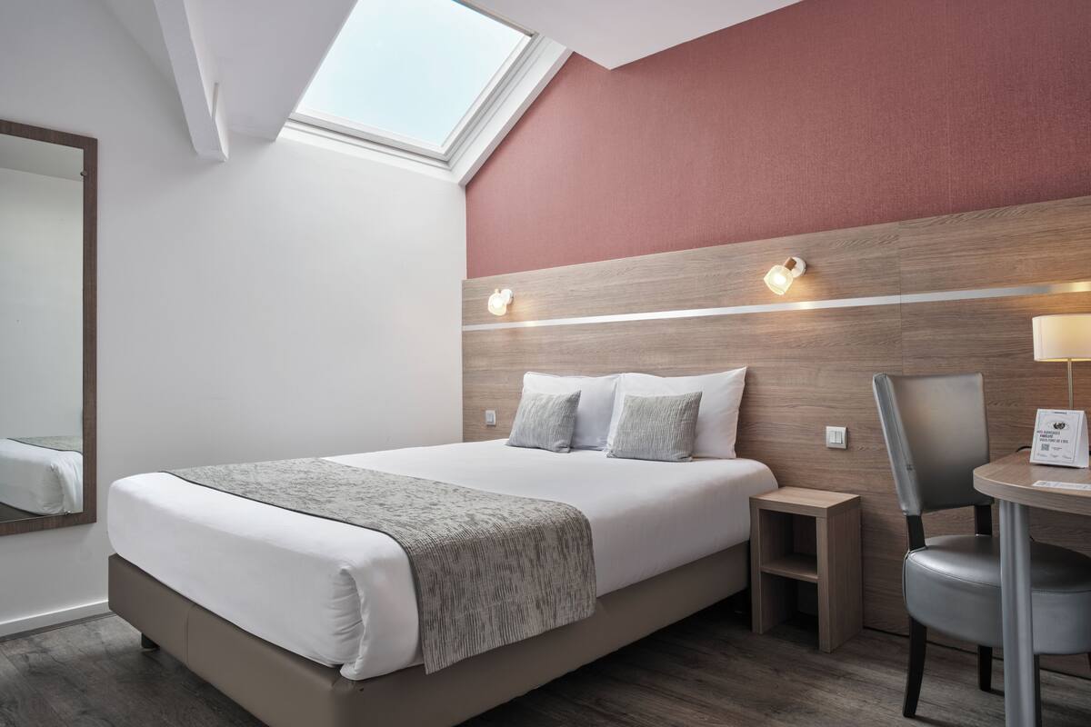 A modern hotel room with a large bed, a small desk, a chair, and a skylight.