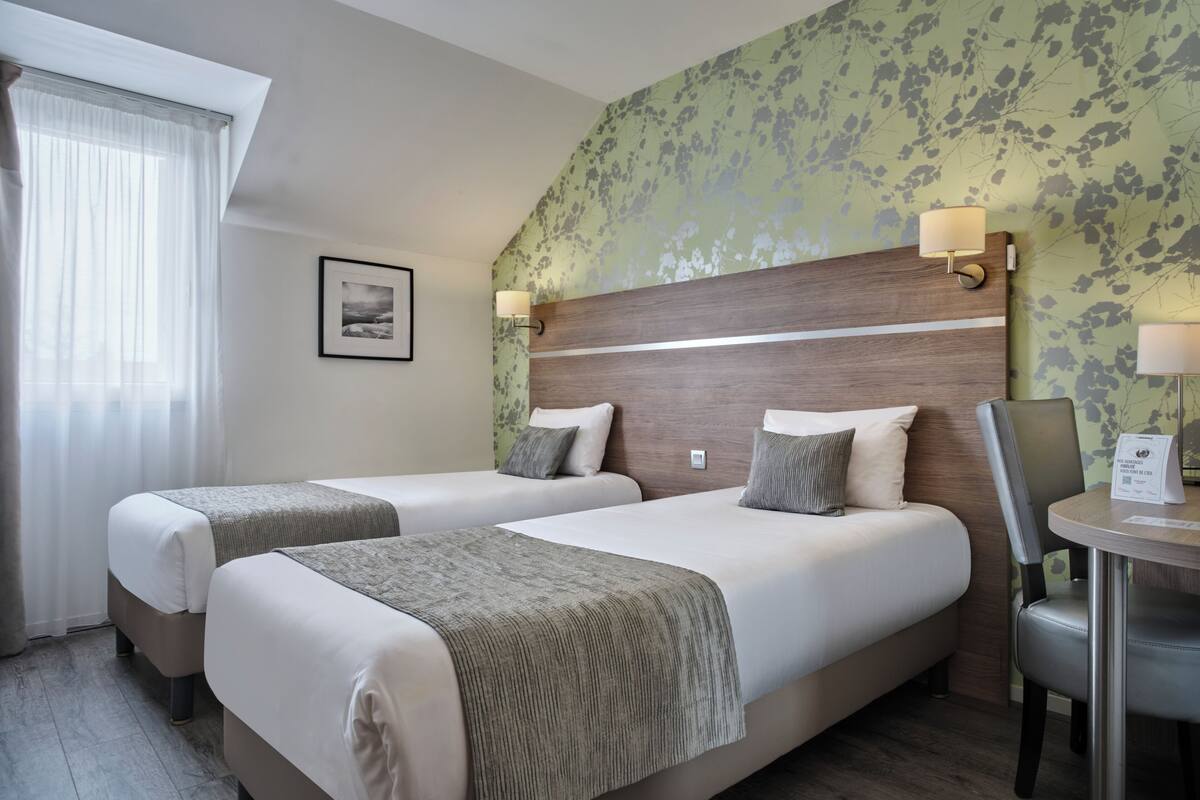 A hotel room with two beds, a wooden headboard, a small table with a lamp, and a picture on the wall.