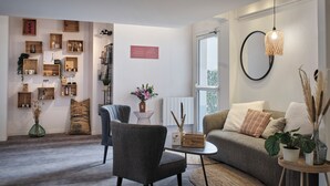 A modern living room with a grey sofa, a coffee table, and a bookshelf. There is a window with a view of the outdoors. - The Originals City, Hôtel Le Beaujoire, Nantes (Nantes)
